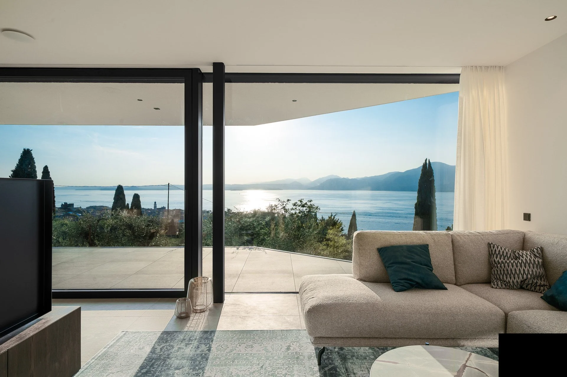 Interno di un soggiorno moderno con divano beige, cuscini blu e neri, vista su un lago con montagne sullo sfondo attraverso grandi vetri scorrevoli.