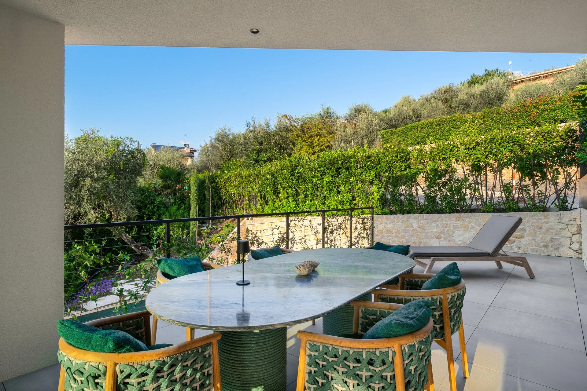Balcone arredato con tavolo e sedie, vista su giardino con cespugli e alberi, sedia a sdraio e cielo azzurro
