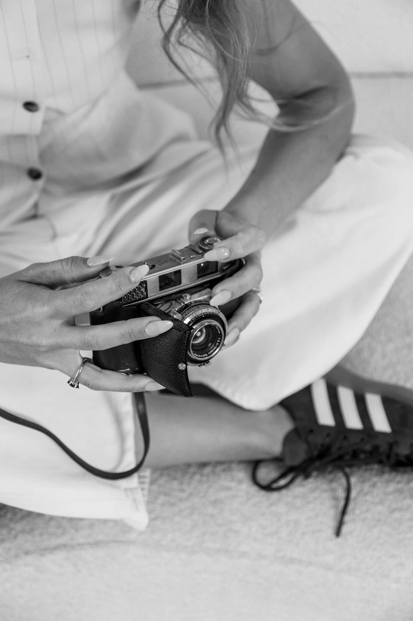 A person sitting on a carpeted floor, holding and examining a vintage camera, wearing a white skirt and a striped shirt.