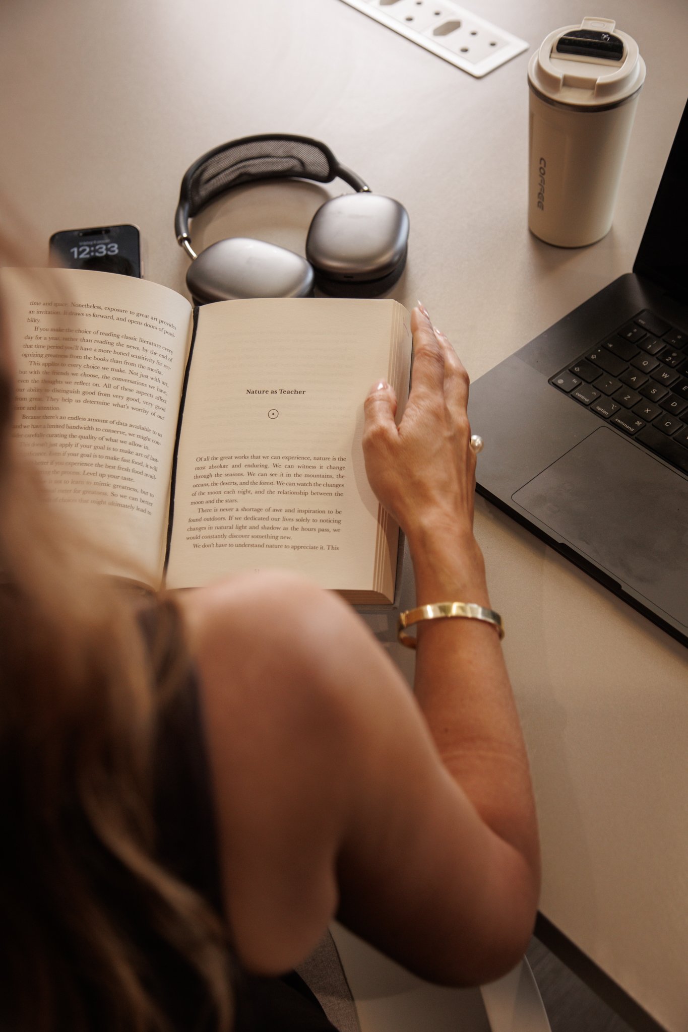 A person reading a book titled 'Nature as Teacher' at a desk with various items including a laptop, smartphone, over-ear headphones, a travel mug, and a wireless charging pad.