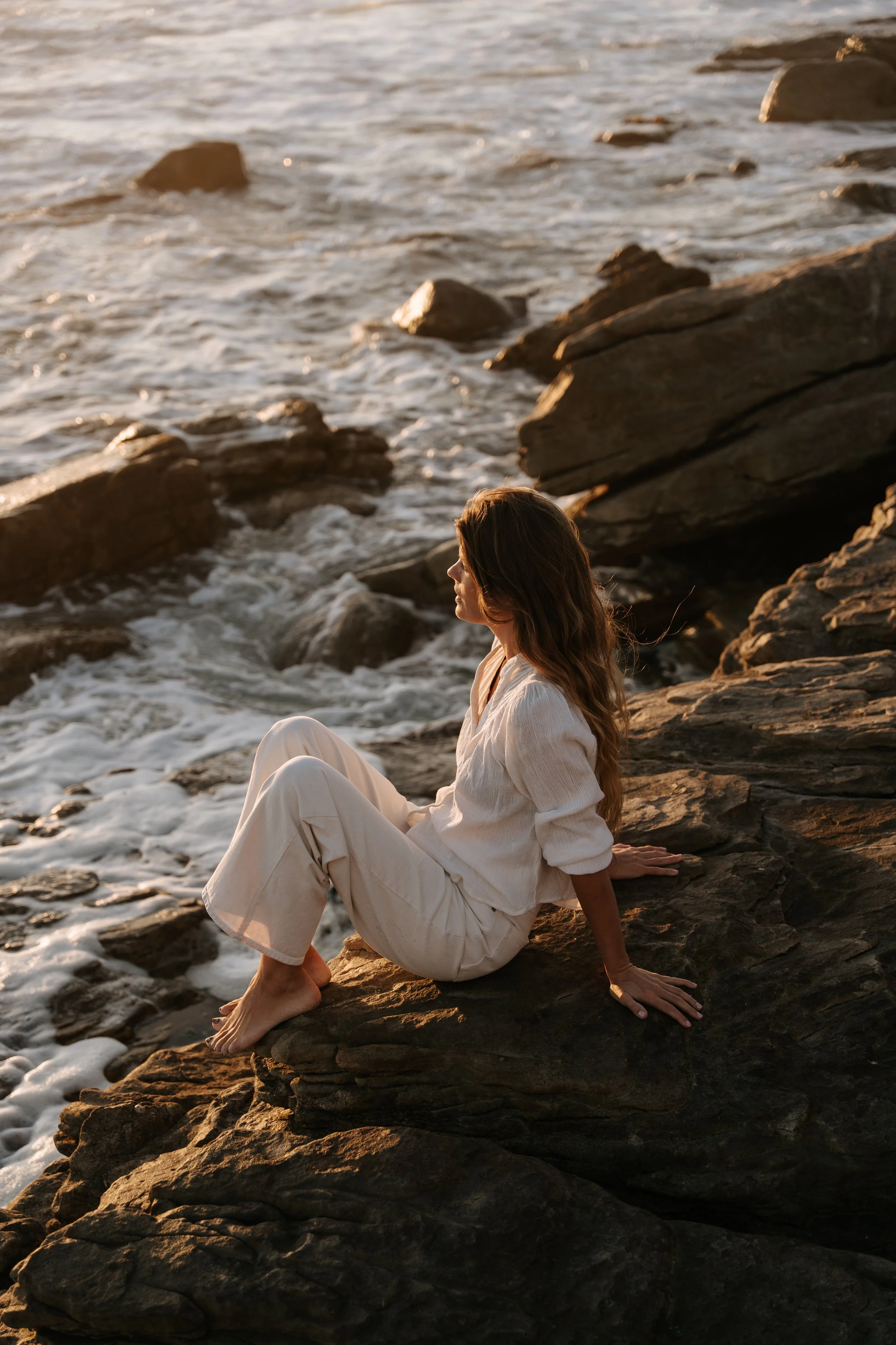 A woman in white clothing sitting on rocks by the ocean during sunset.