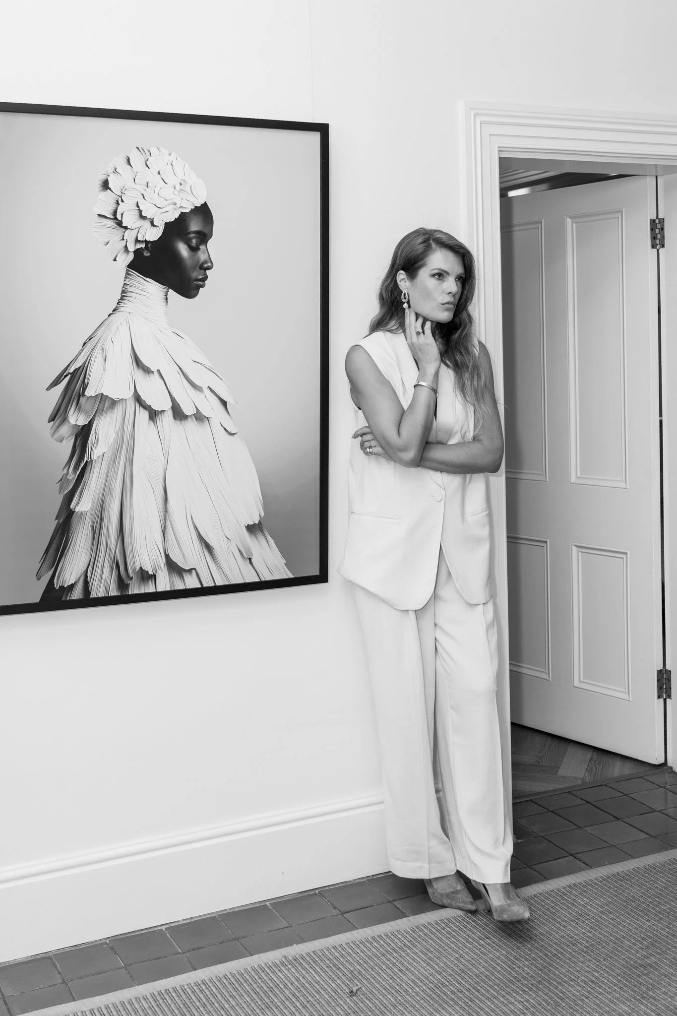 A woman in a white suit stands with her arms crossed, touching her face, next to a large black and white portrait of a woman with elaborate feathered headwear and layered feathered clothing.