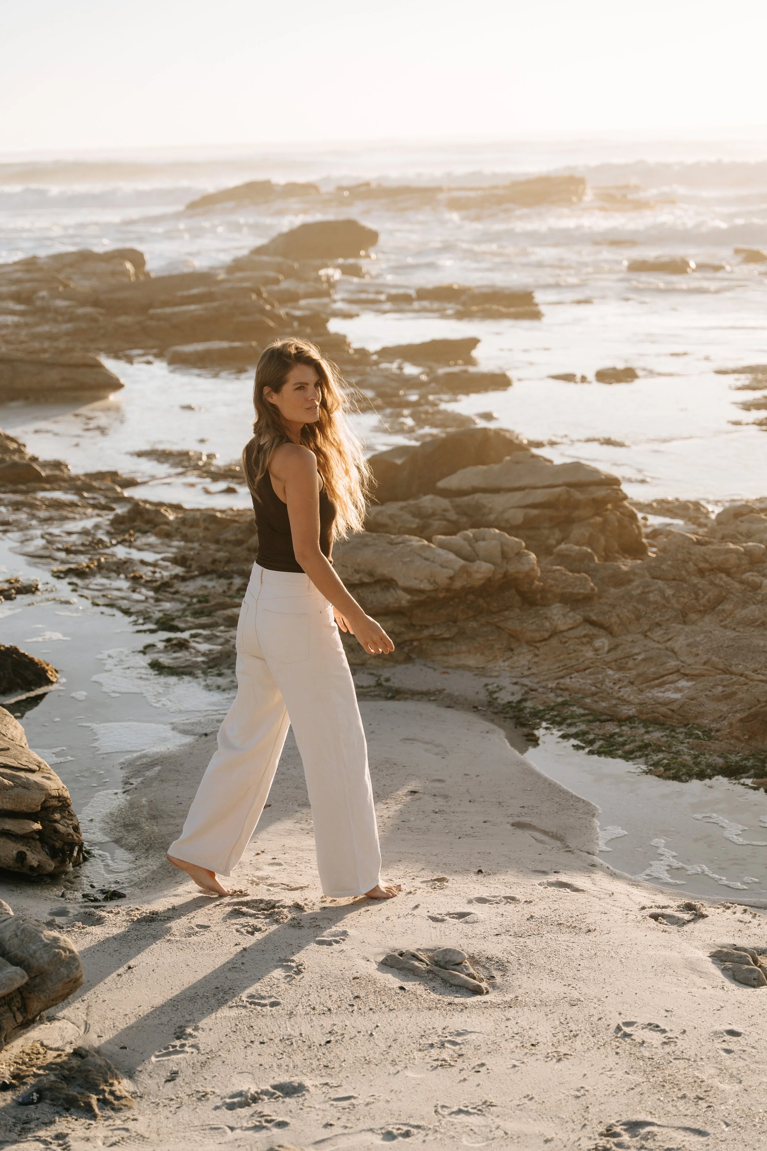 A woman walking barefoot along a sandy beach with rocks, during sunset, with ocean waves in the background.