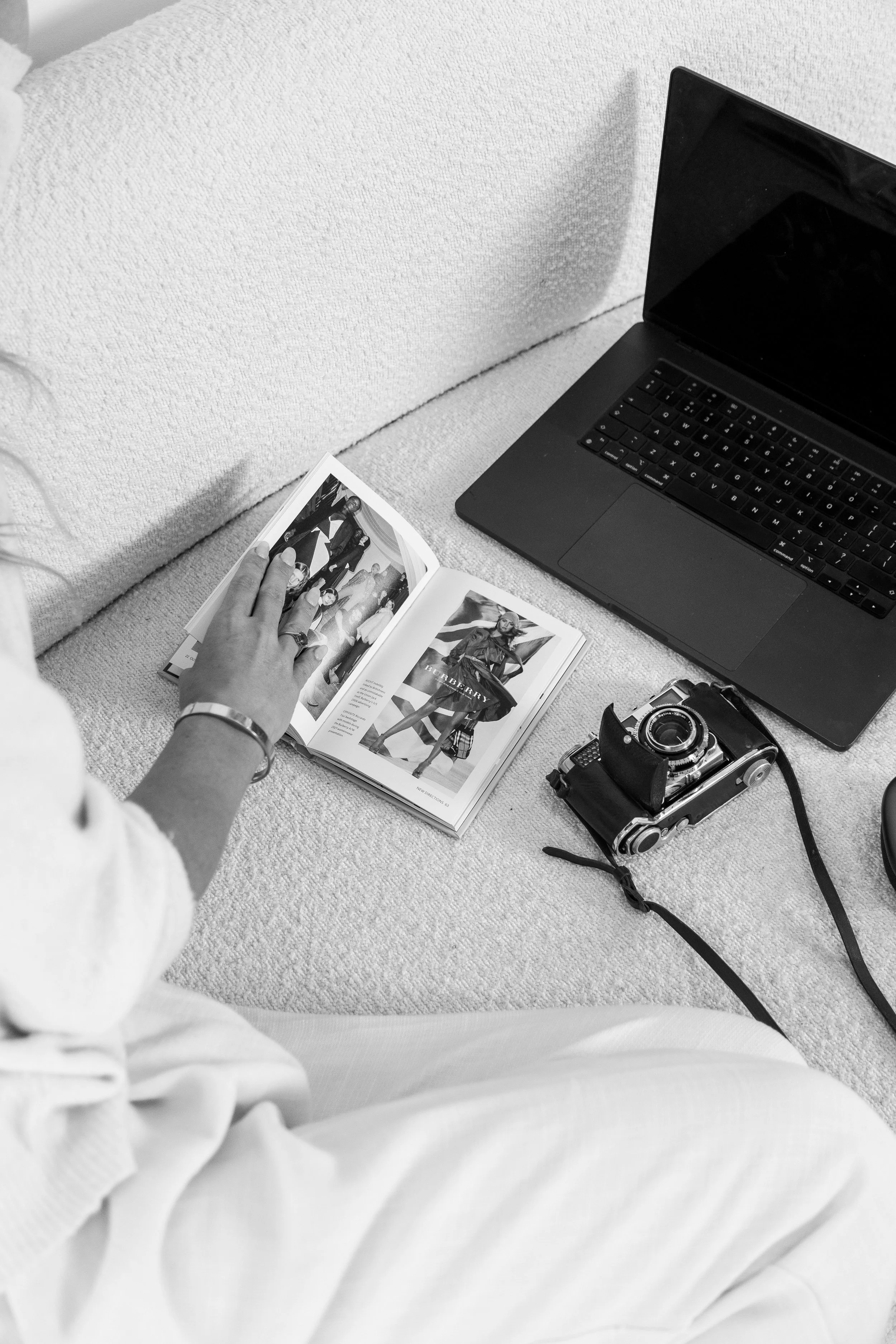 Person sitting on a sofa, looking at a magazine with fashion photos. A camera, laptop, and part of the sofa are also visible.