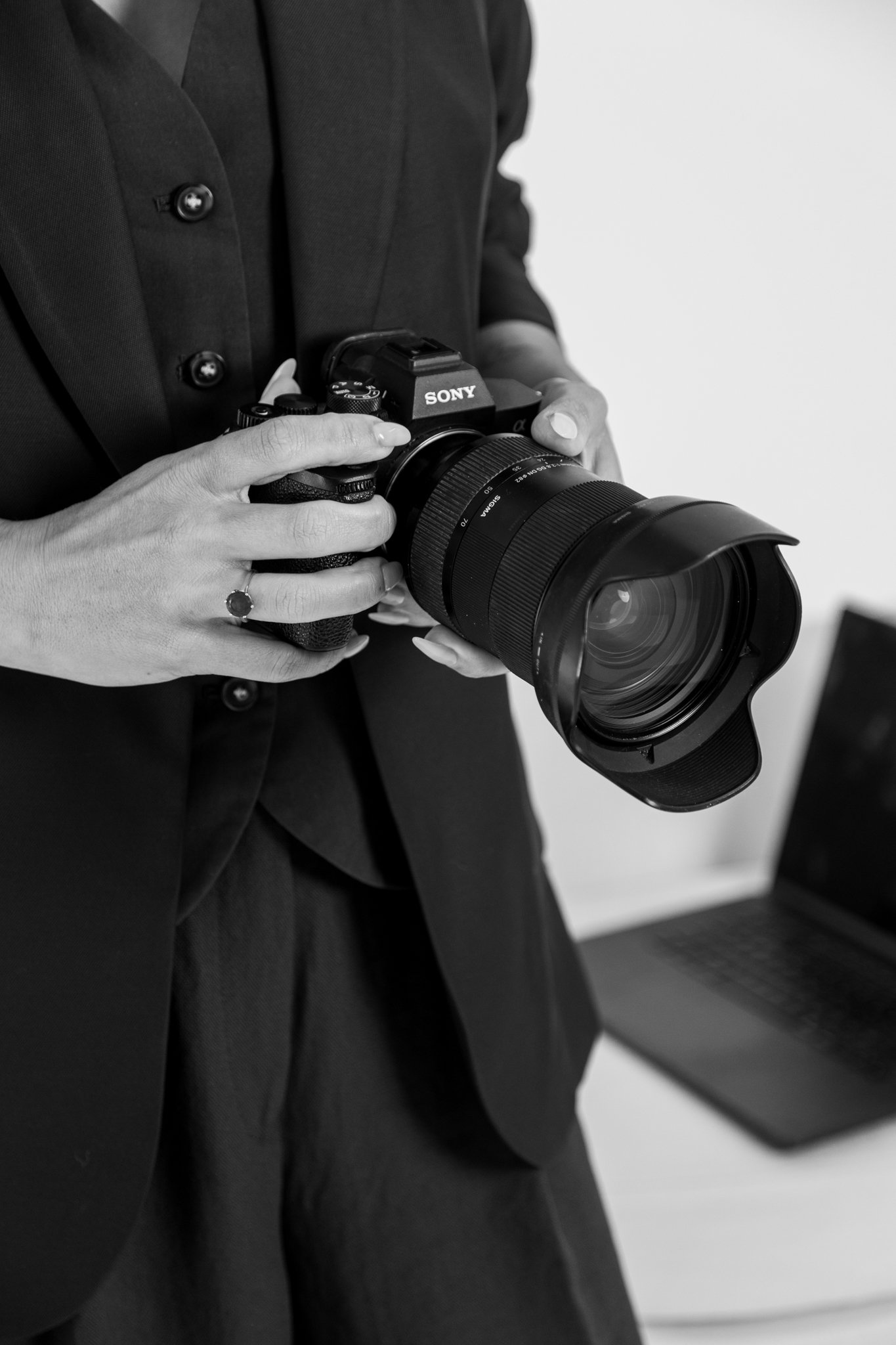 A person in a dark jacket holding a professional Sony camera with a large lens, standing near a laptop.