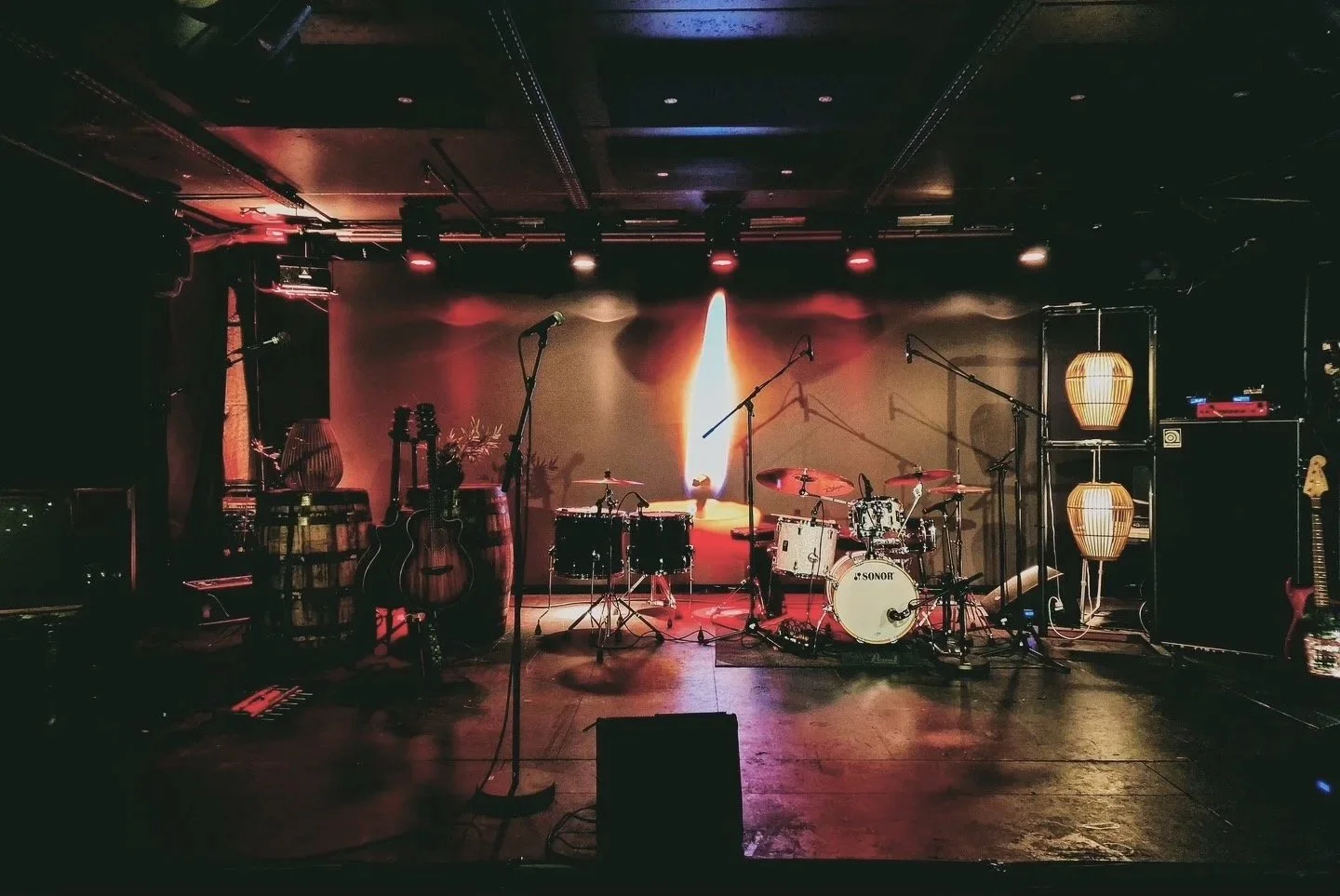 Scene med musikkinstrumenter oppstilt, inkludert trommer, gitarer, mikrofoner og forsterkere, opplyst av varme scenelys, med mørk bakgrunn og koselige lysarmaturer.