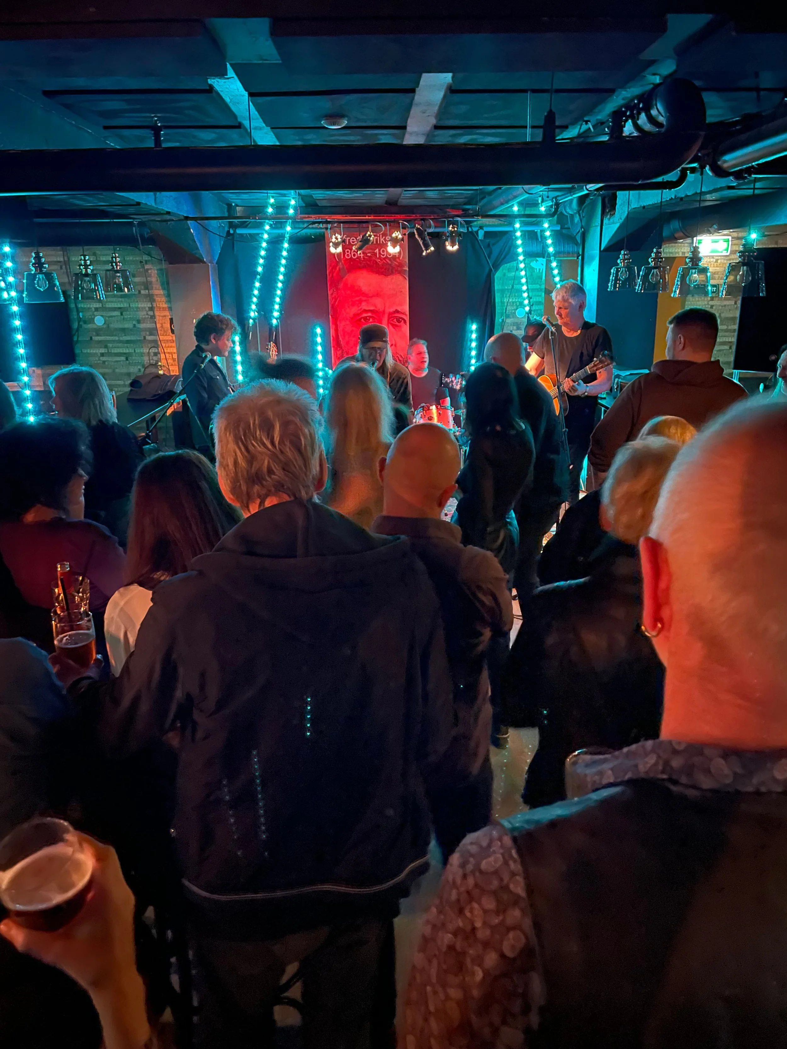 Live musikkopptreden i et svakt opplyst lokale med en folkemengde samlet foran en scene, et band som spiller gitarer og trommer, med fargerike lys og en rød plakat i bakgrunnen.