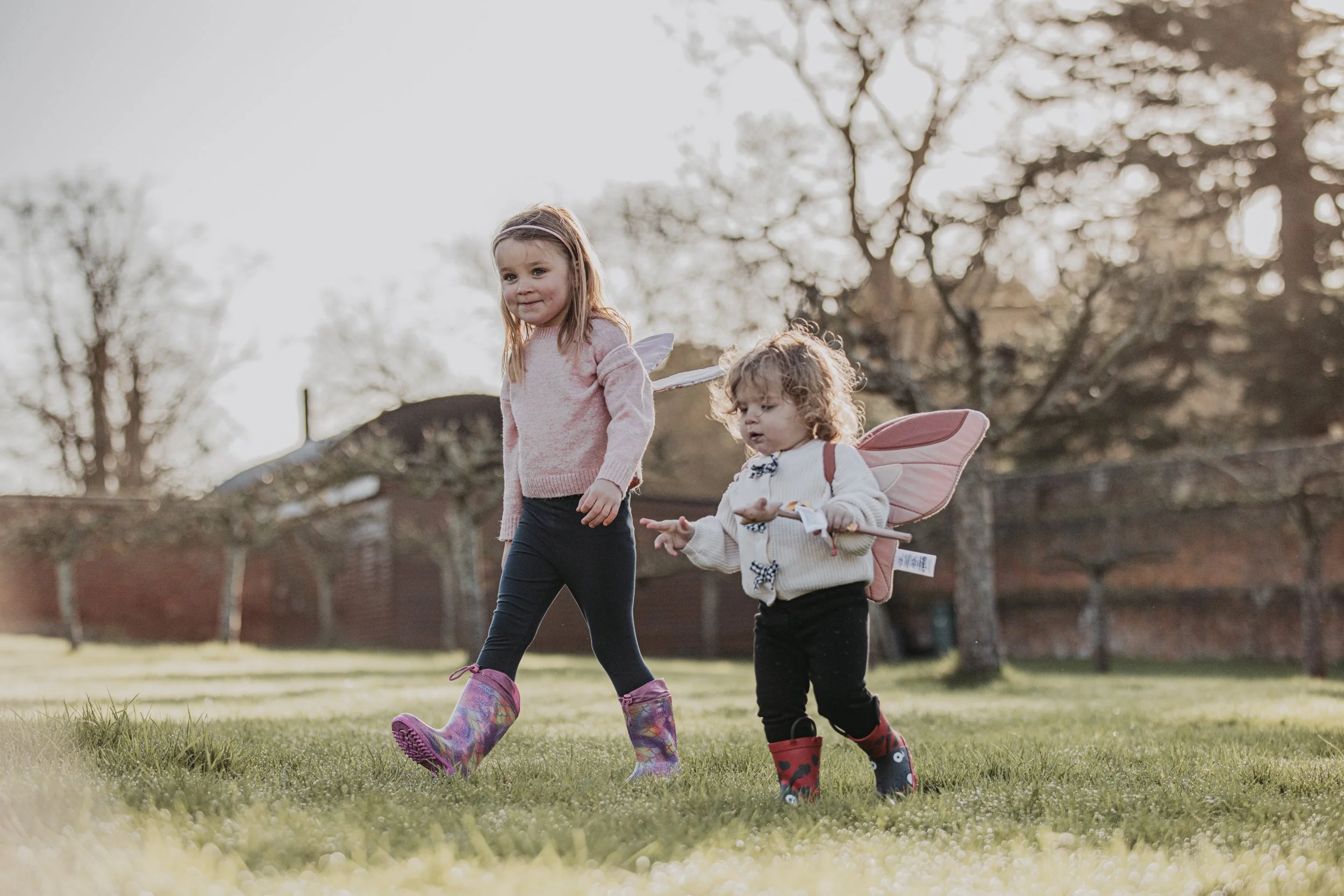 Two young girls, dressed as fairies with wings, riding on grass in a park during daytime. One girl has brown hair, a pink sweater, and black leggings, wearing colorful rain boots. The other girl has curly light brown hair, a white sweater, and black pants, with red and black rain boots. Trees and a brick wall are visible in the background.