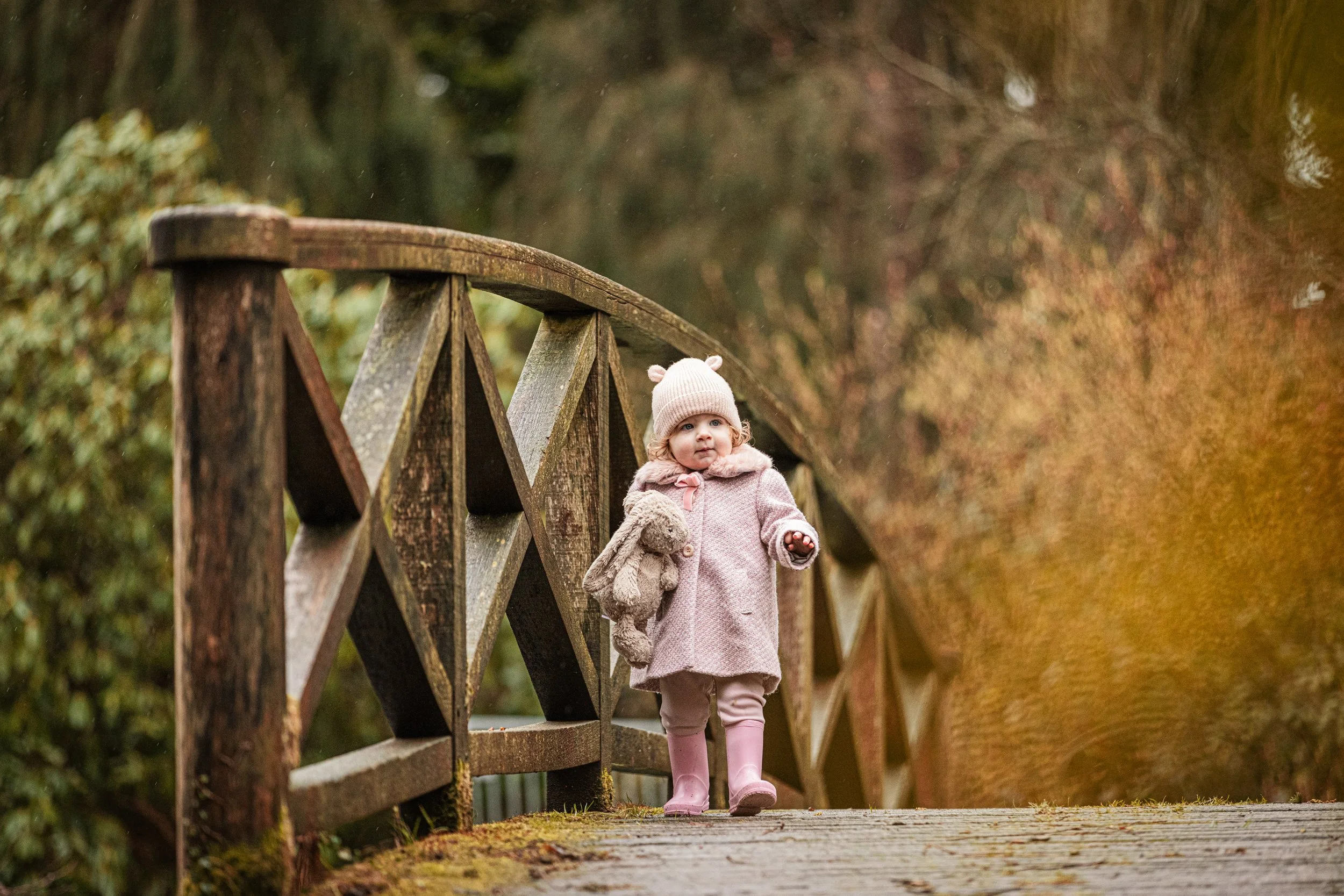 A young girl in a pink coat, hat, and leggings walking on a wooden bridge with a stuffed animal in hand, surrounded by autumn trees.