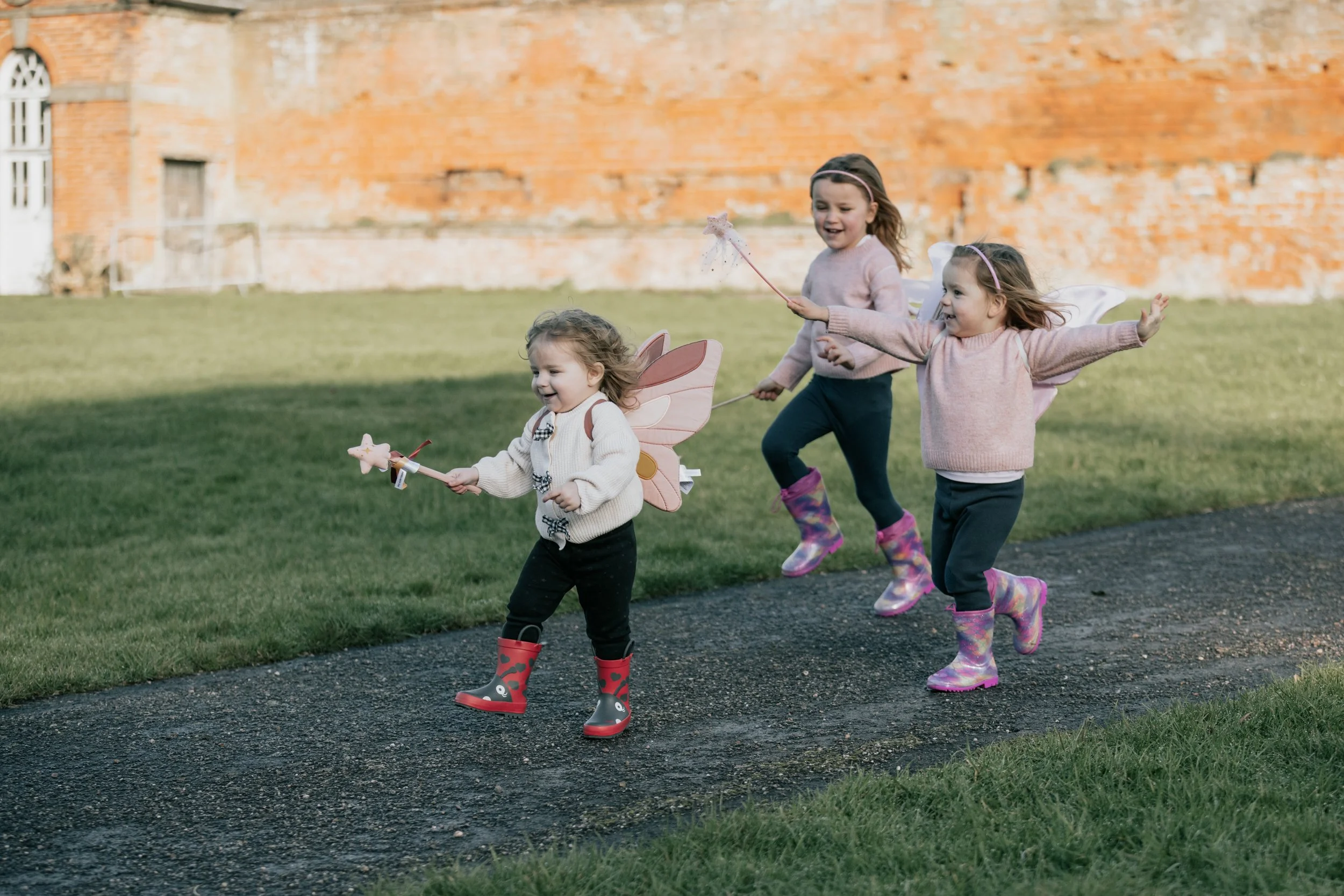Three young girls running on a paved path, dressed in fairy costumes with wings and holding wands, with a brick building in the background.