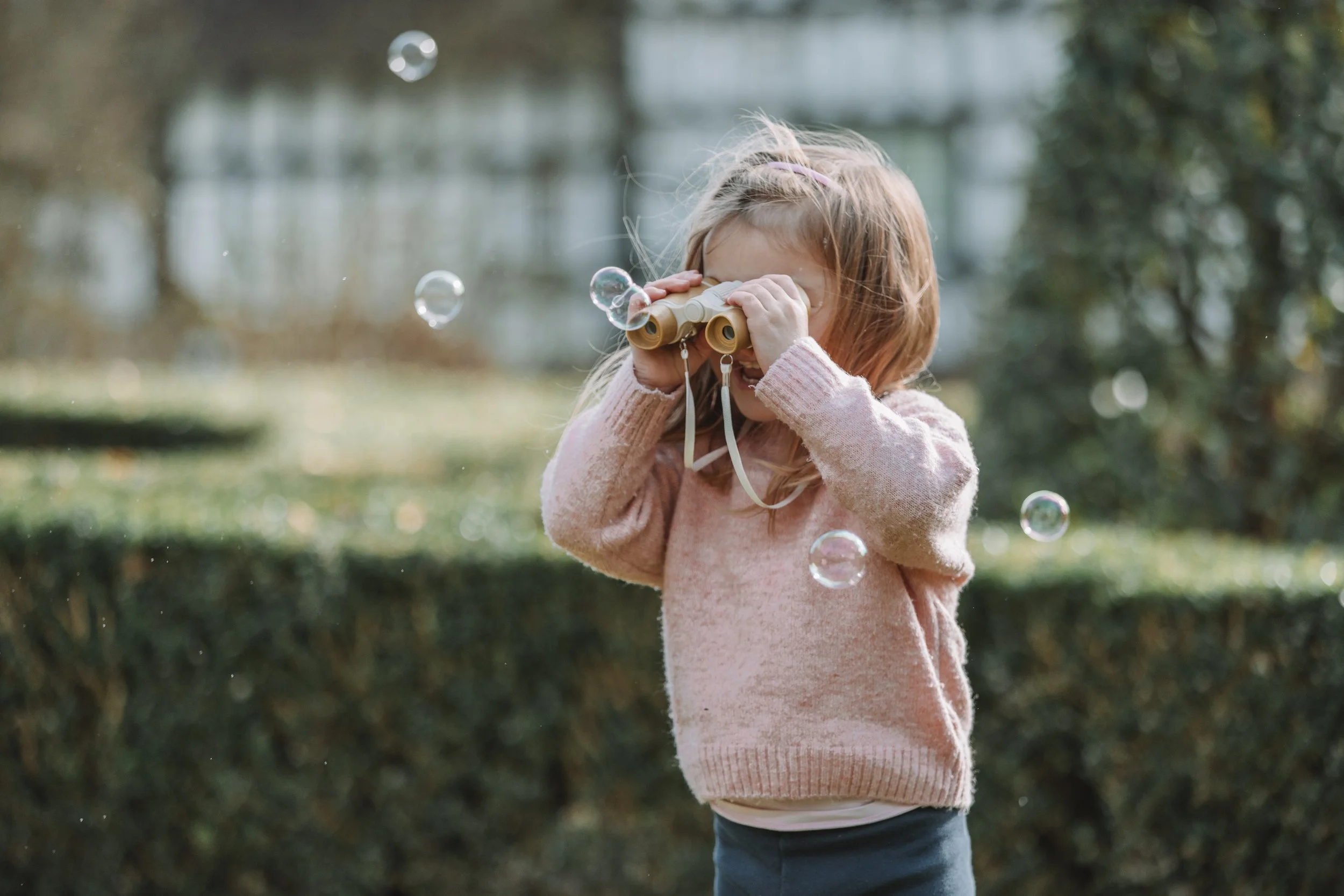 A young girl with long blonde hair looking through binoculars in an outdoor park, surrounded by floating soap bubbles.