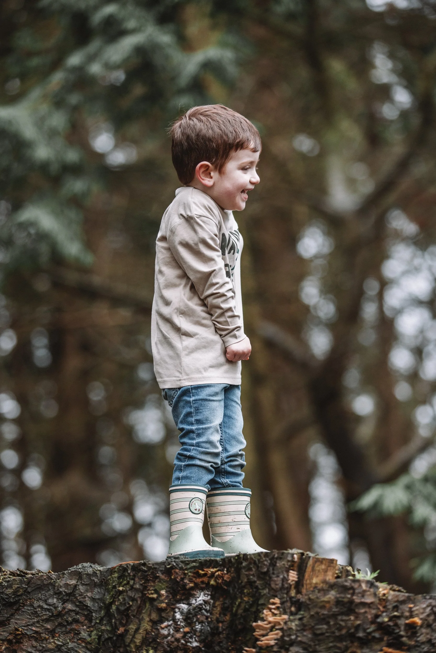 Young boy standing on a tree stump outdoors, wearing rain boots and smiling.