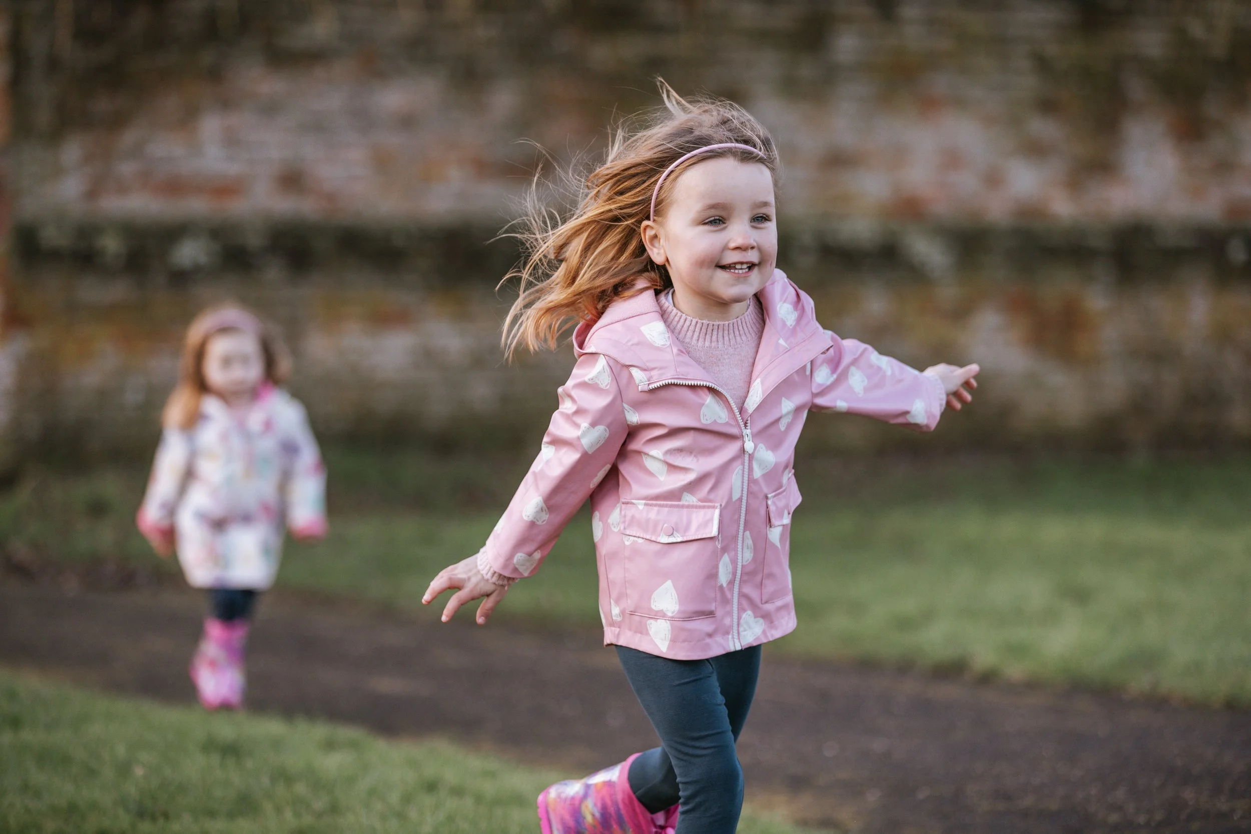 Two young girls with red hair wearing pink jackets playing outside near a pond, with grass and a brick wall in the background.