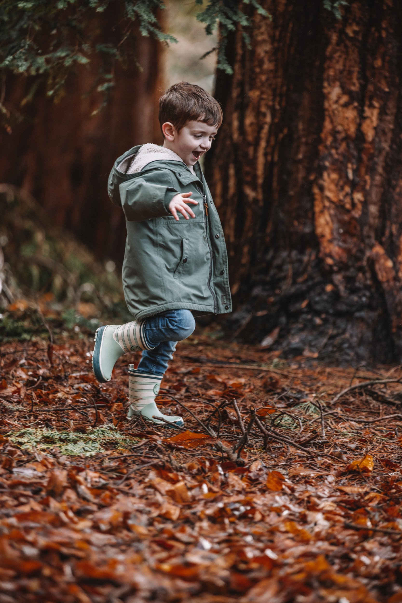 A young boy in a green jacket and striped rain boots is outdoors on a forest floor covered with autumn leaves, playing or jumping near a large tree.