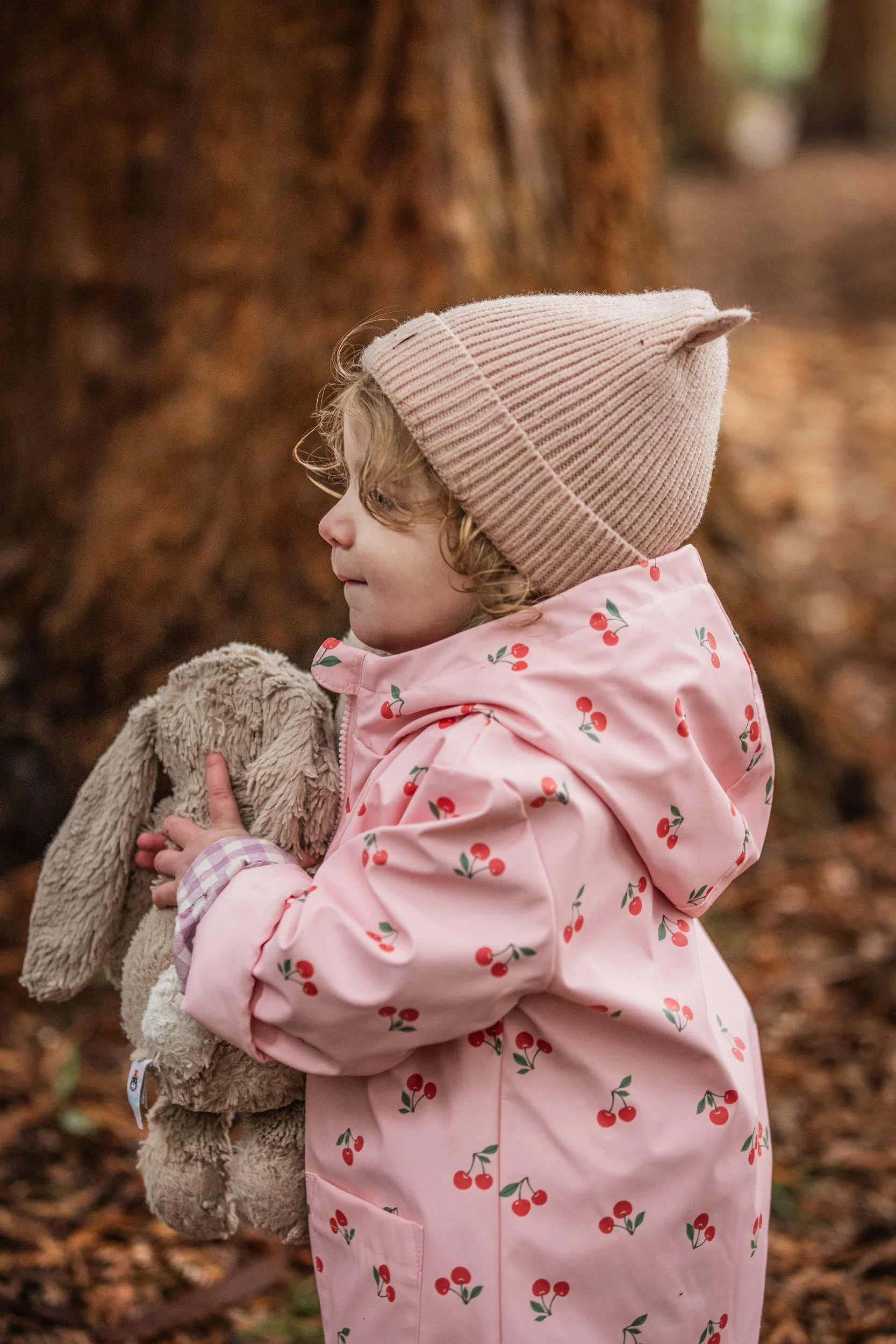 A young girl with curly blonde hair, wearing a pink coat with red cherries, a beige knit hat, and holding a plush bunny, standing outdoors in a forested area with autumn leaves on the ground.