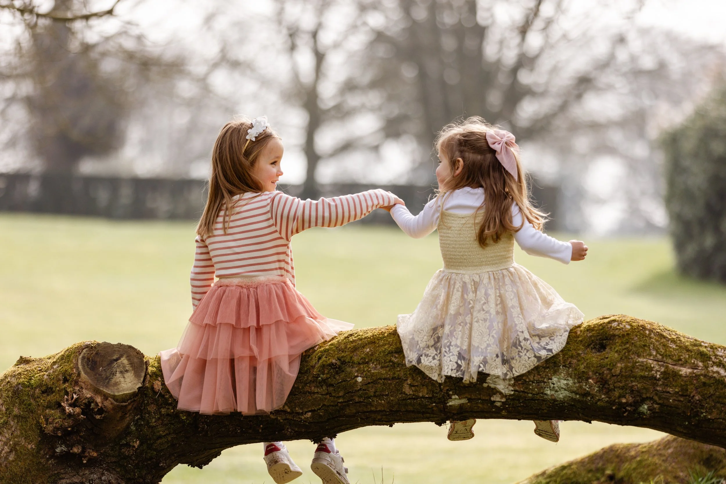 Two young girls sitting on a mossy tree trunk in a park, holding hands, and smiling at each other, with blurred trees and grass in the background.