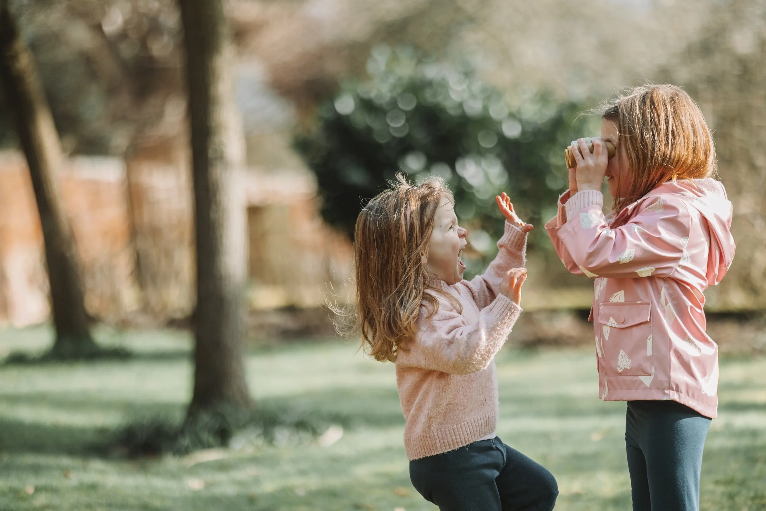 Two young girls playing outdoors in a park, one is shouting while the other is looking through a binoculars.