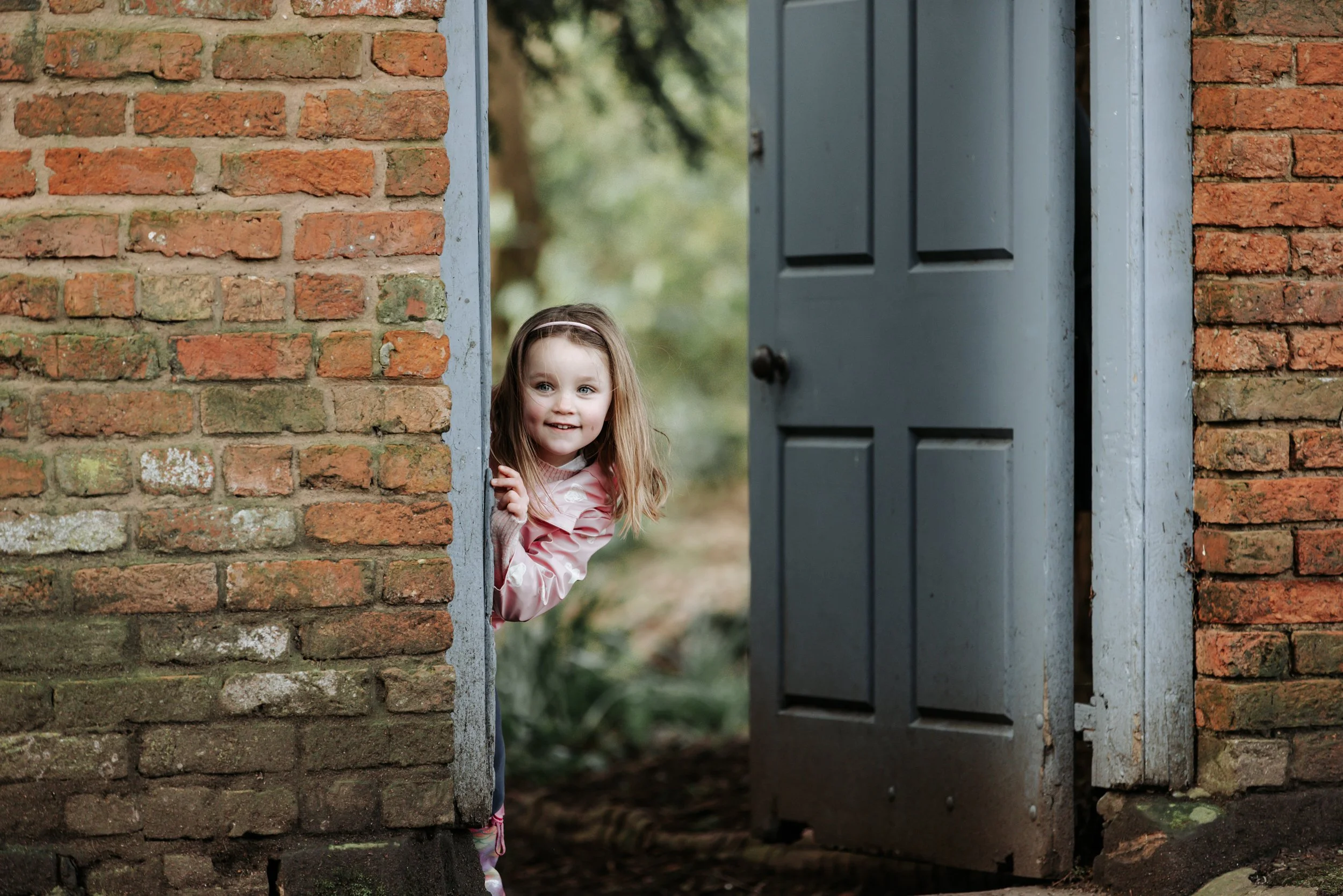 A young girl with long brown hair and a pink shirt peeks out from behind a brick wall, smiling. She is standing near a weathered blue door that is slightly open, revealing greenery in the background.