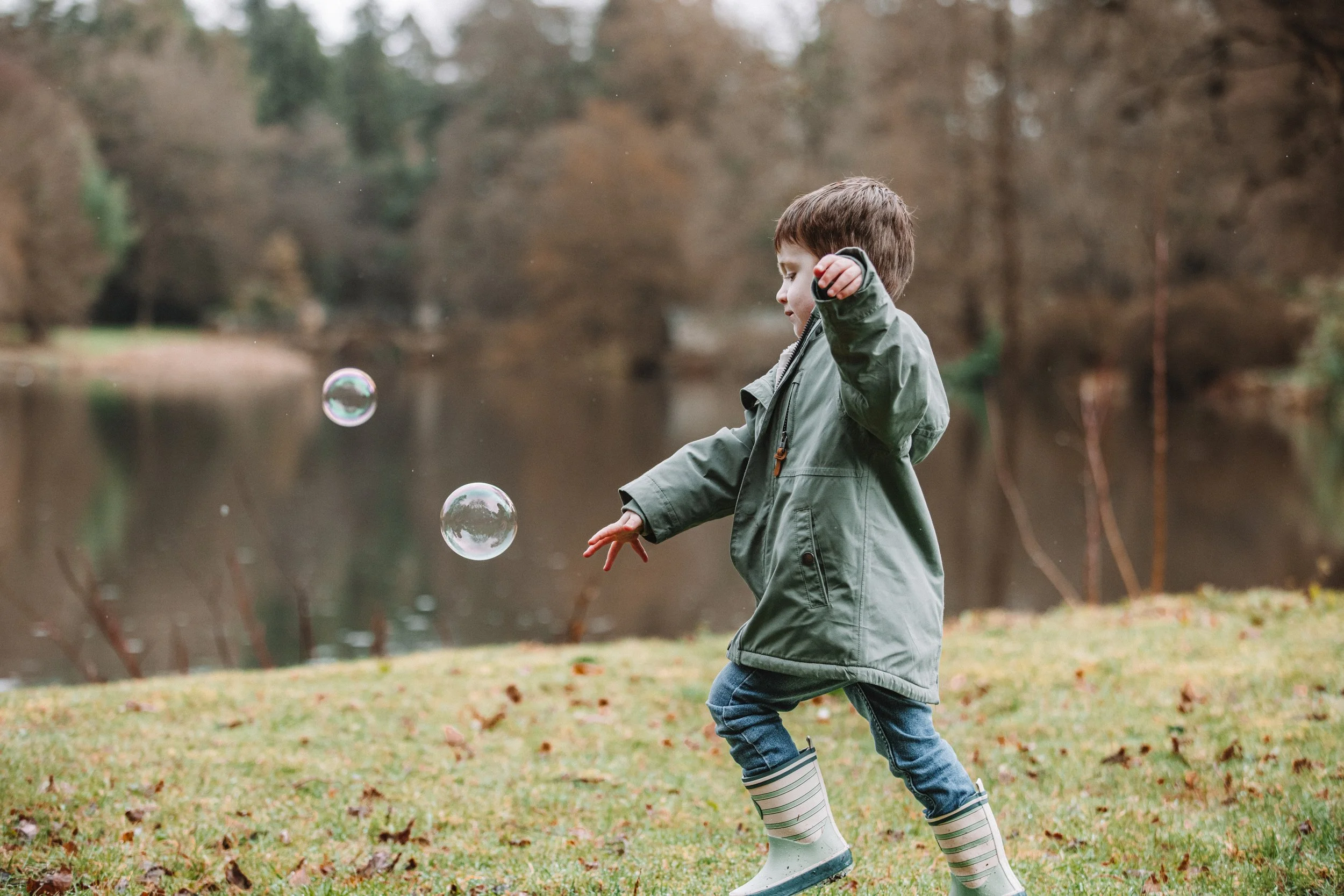 A young boy in a green raincoat and striped rain boots plays with soap bubbles by a lake or pond in an outdoor park during fall.
