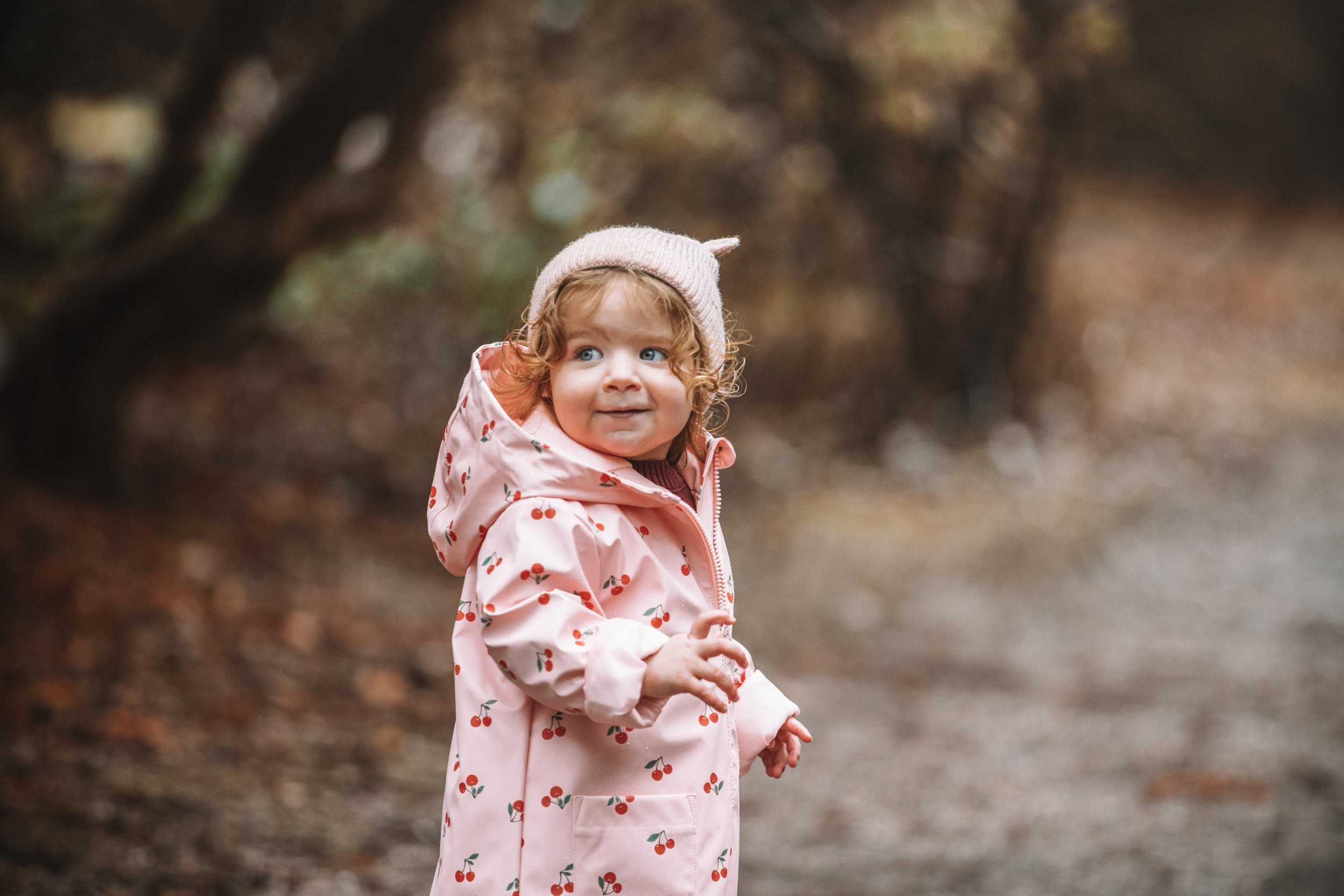 A young girl with curly hair wearing a pink cherry-print raincoat and a knit hat, standing outdoors in a wooded area with autumn-colored leaves.