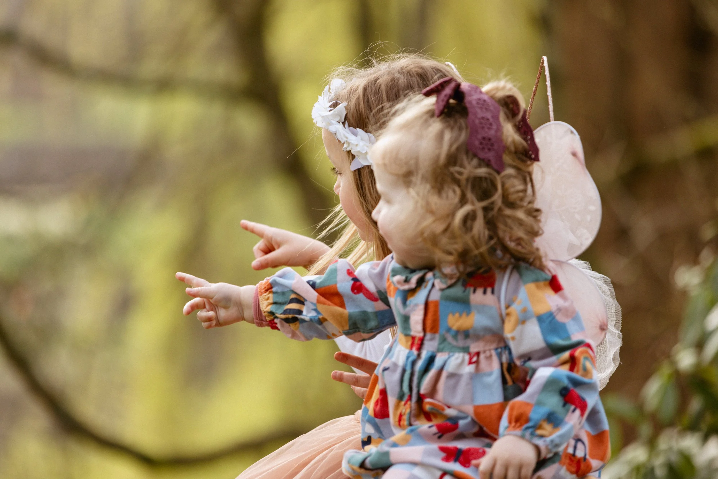 Two young girls in colorful outfits and floral headbands sitting outdoors on a tree branch, pointing at something off in the distance.