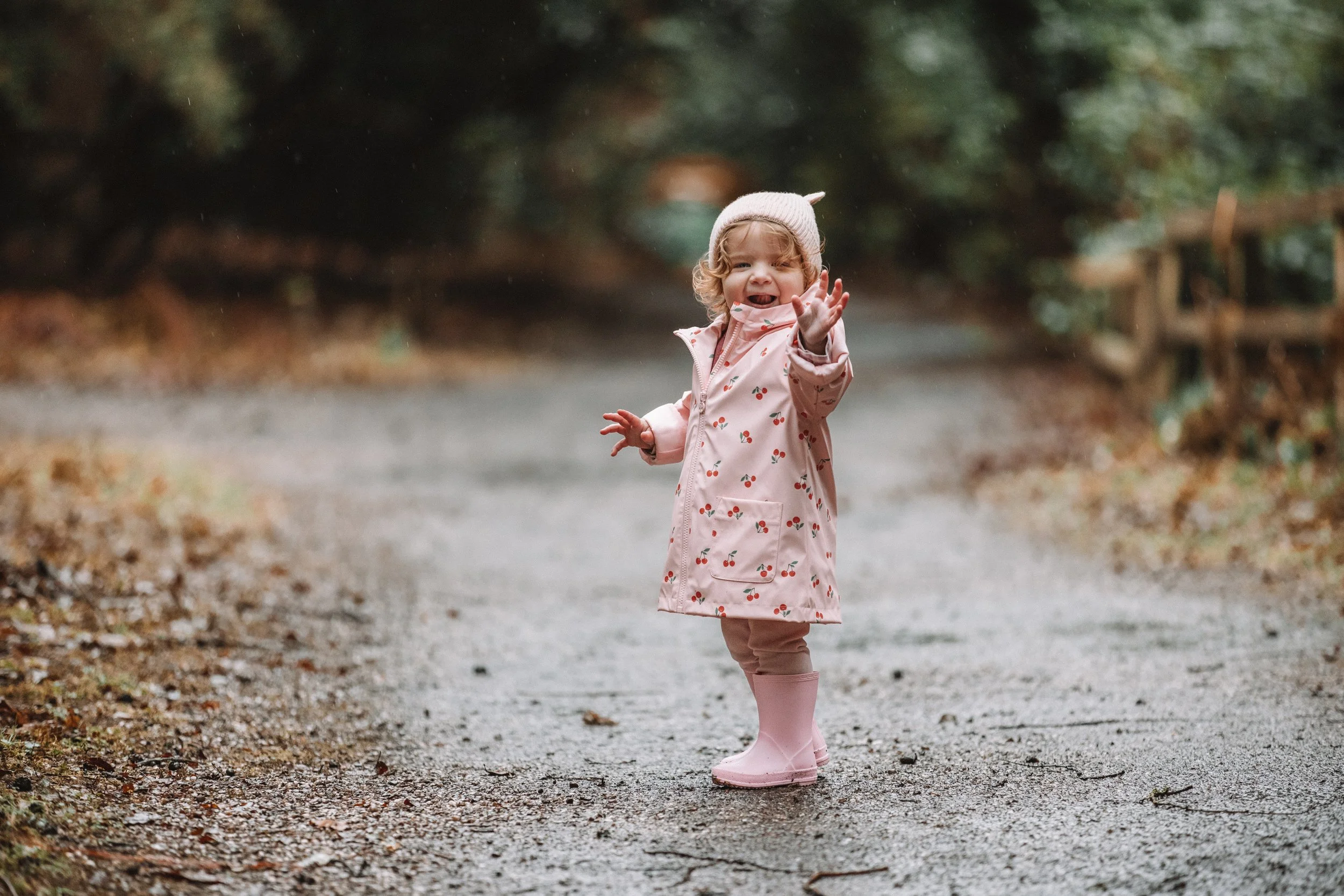 A young girl with curly hair, wearing a pink raincoat with cherry patterns, pink rain boots, and a white knit hat, waving and smiling on a dirt path outdoors.