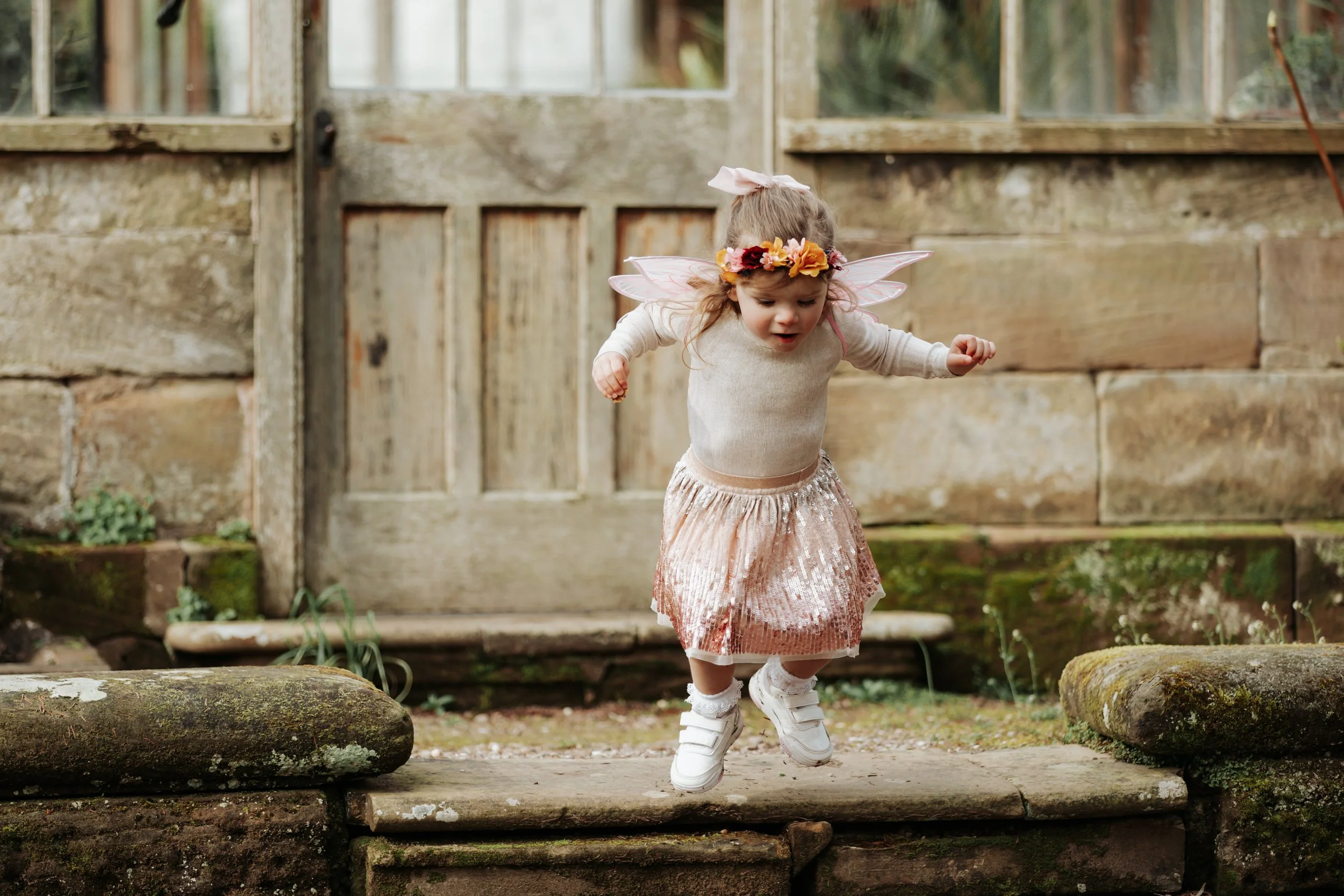 A young girl dressed as a fairy jumps off a stone step outdoors. She is wearing a pink, sequined skirt, a beige top, white sneakers, fairy wings, and a floral headband and bracelet.
