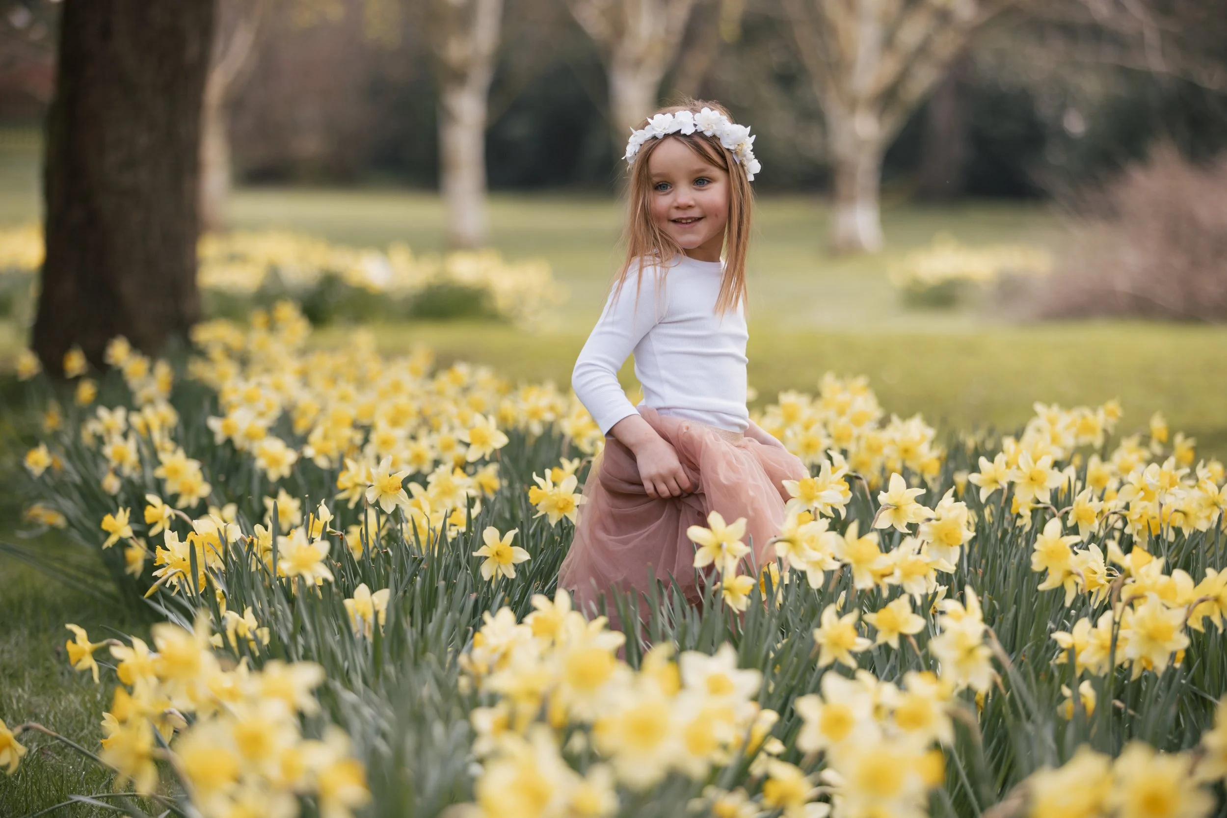 A young girl with a flower crown smiling and holding her skirt while standing among yellow daffodils in a park.