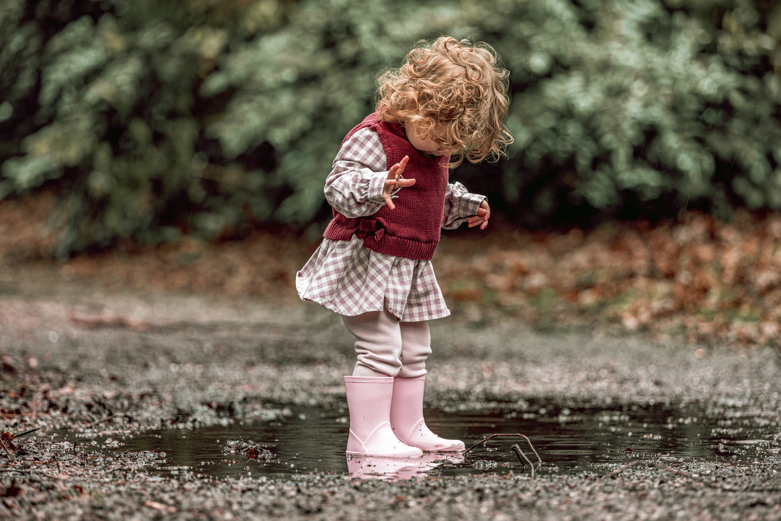 A young girl with curly hair in a checkered dress, pink rain boots, and a maroon sweater vest standing in a puddle outdoors.