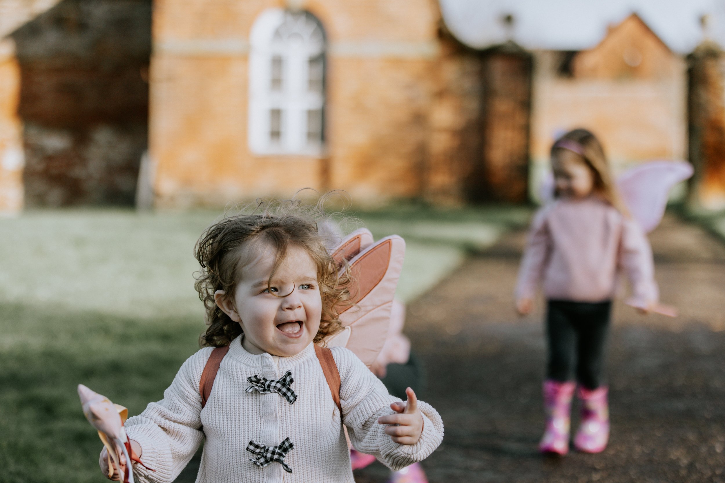 Two young girls with fairy wings playing outdoors near a brick building.