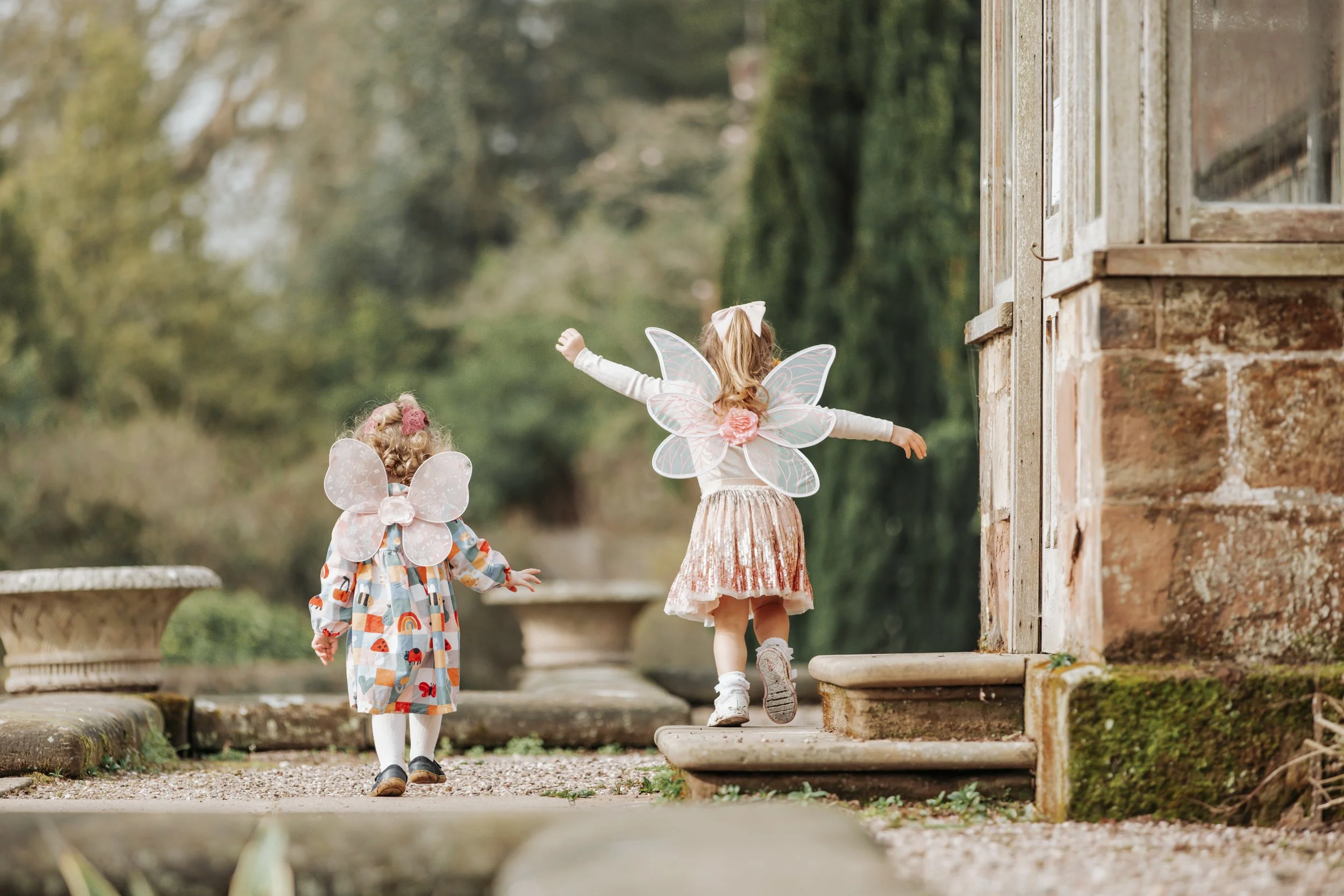 Two young girls dressed as fairies with wings, walking outdoors near a rustic brick building with steps, surrounded by trees.