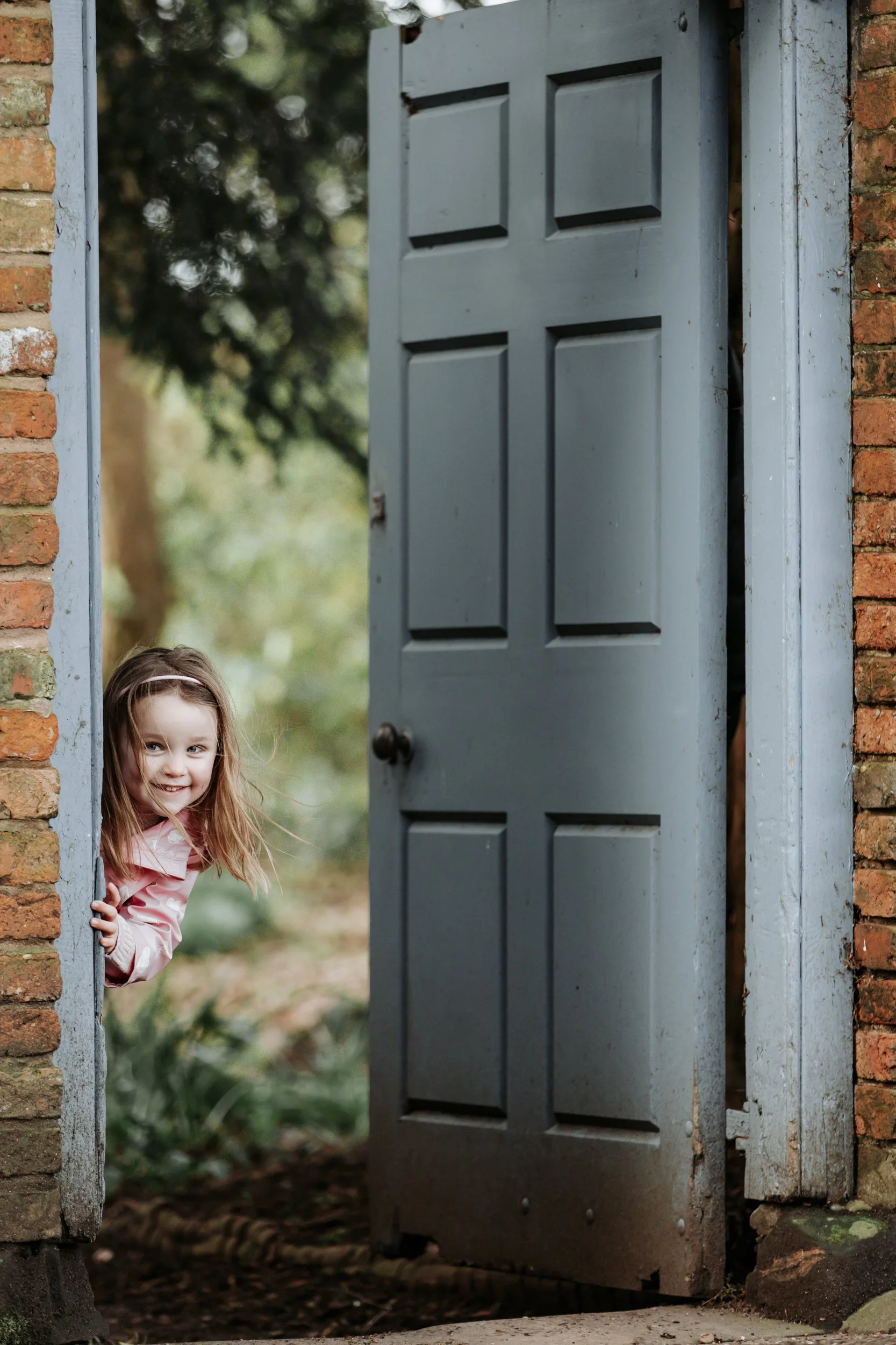 A little girl with light brown hair peeking out from behind an open blue door, smiling and looking at the camera.