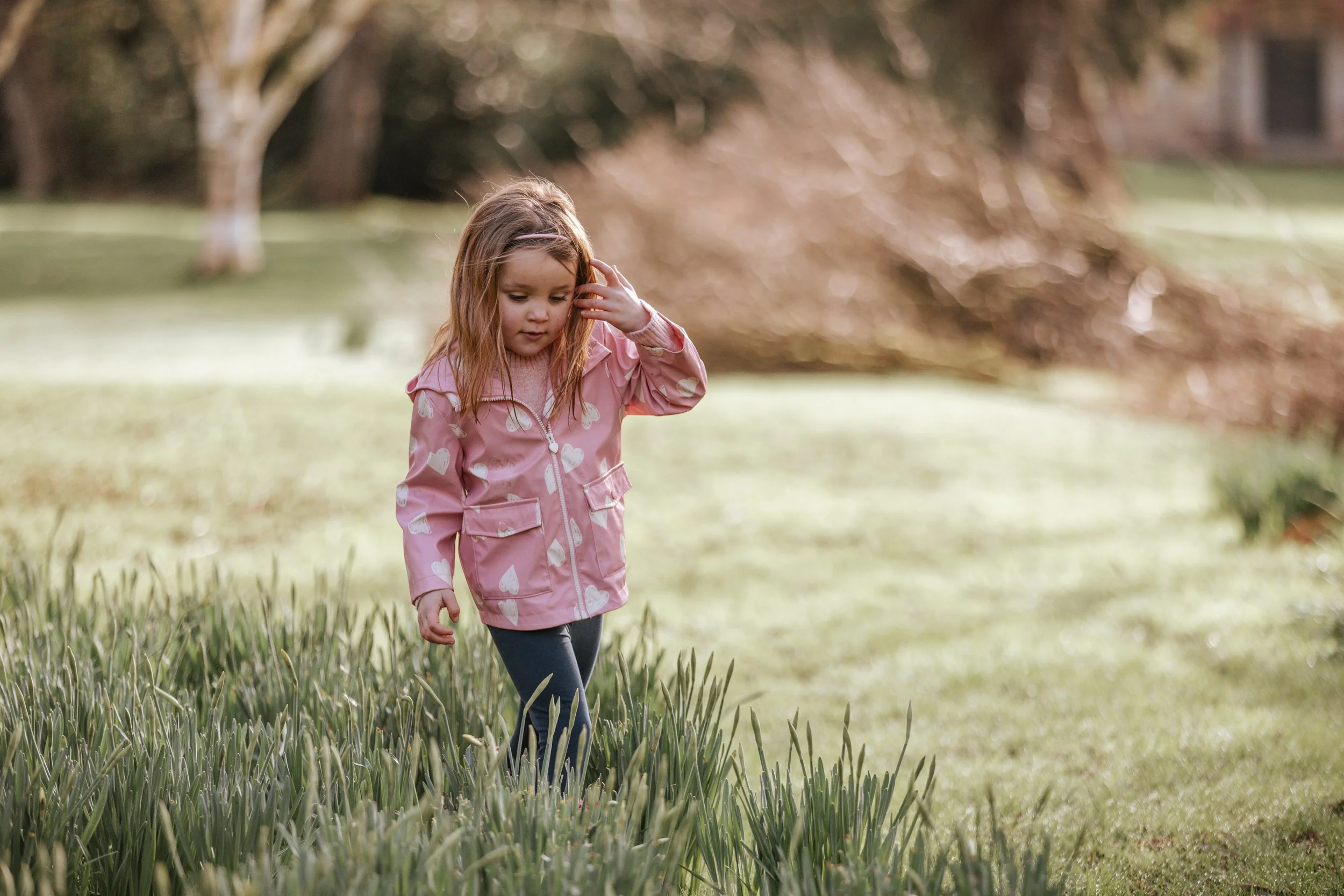 A young girl with long, red hair, wearing a pink jacket with white hearts, walking through a grassy area outdoors on a sunny day.