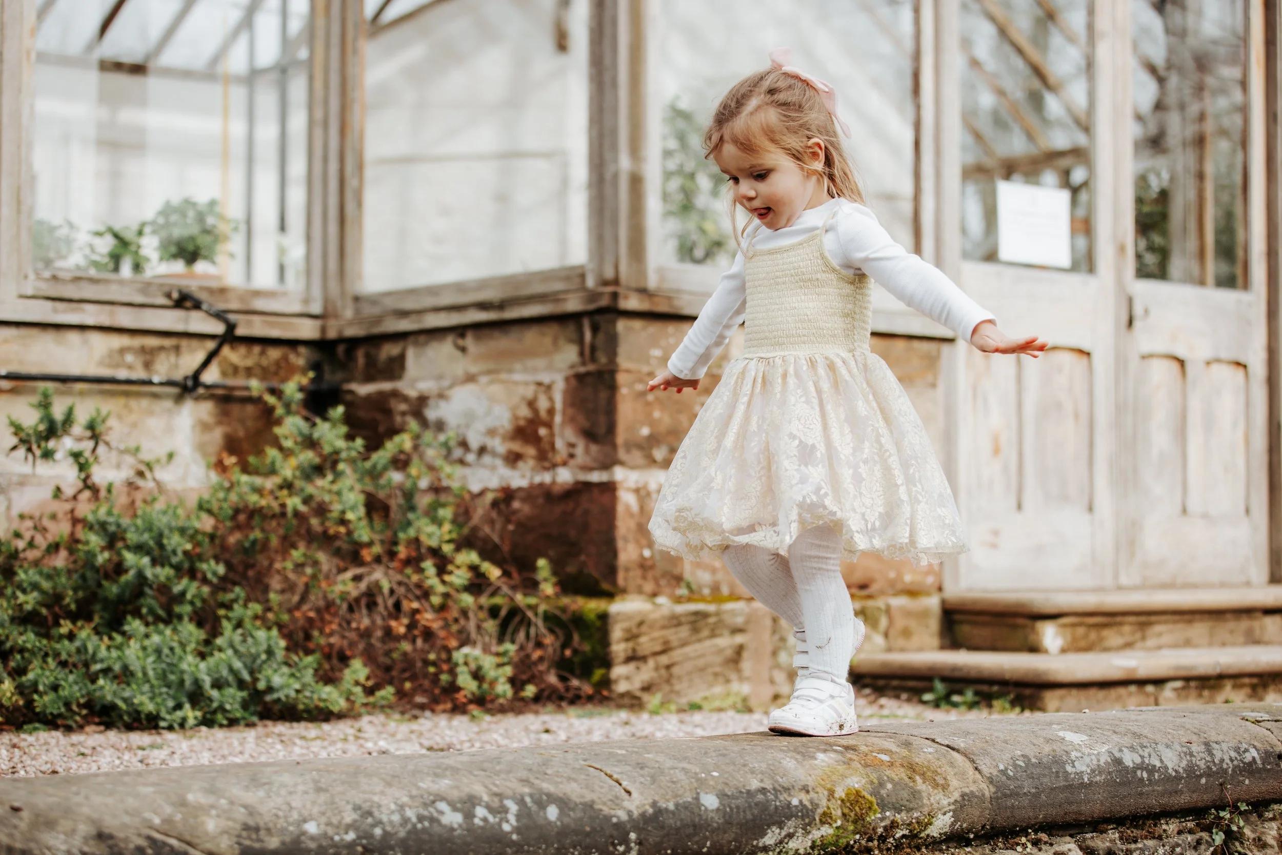 A young girl in a cream-colored dress and white sneakers balancing on a stone edge outside a rustic glass greenhouse.