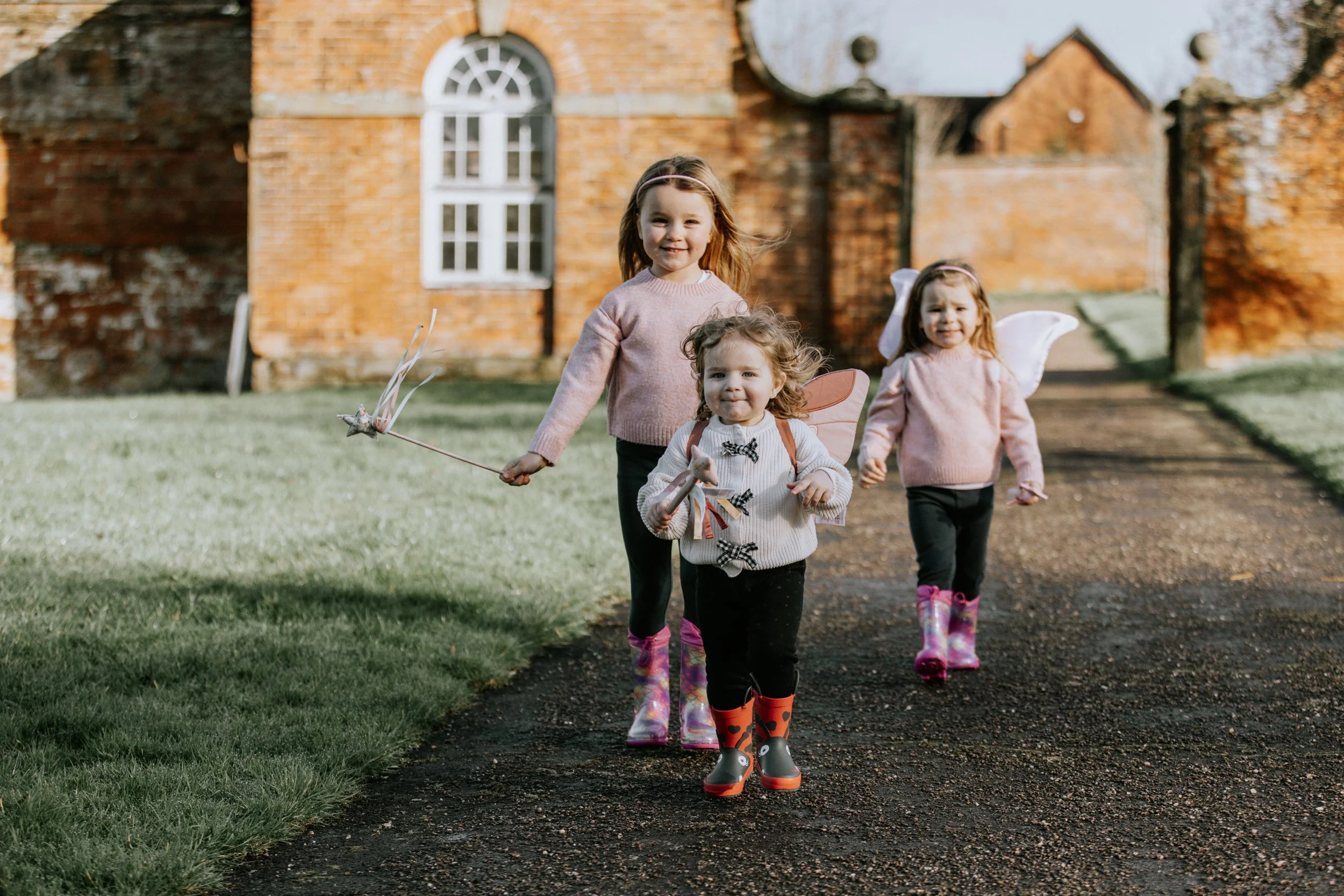 Three young girls with fairy wings and holding fairy wands walk along a paved path outside in front of a brick building, dressed in pink sweaters and rain boots, smiling and enjoying a nature walk.