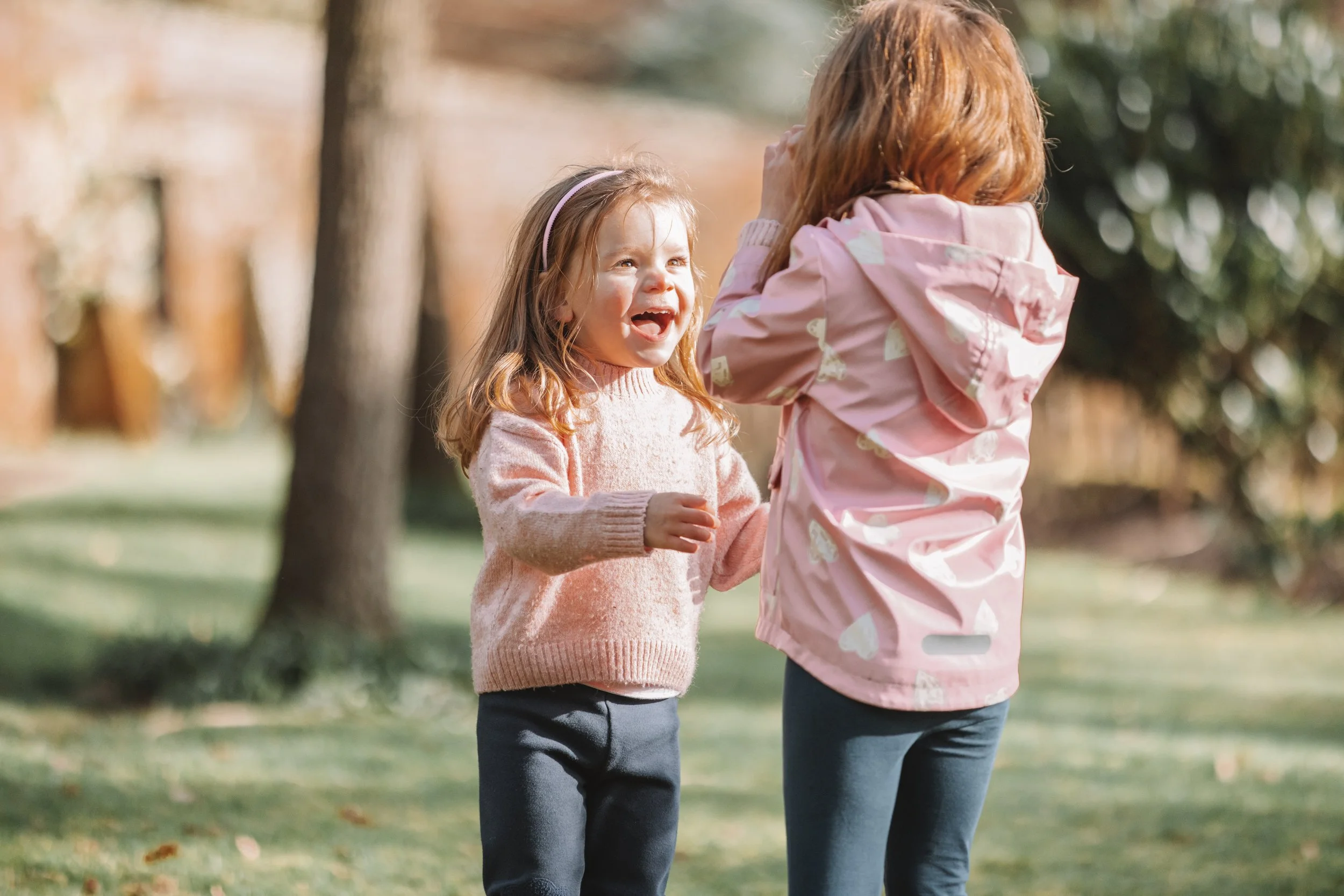 Two young girls outdoors in a park, one with curly hair in a pink sweater smiling and laughing, the other with straight hair in a pink raincoat with white pattern, covering her face with her hands.