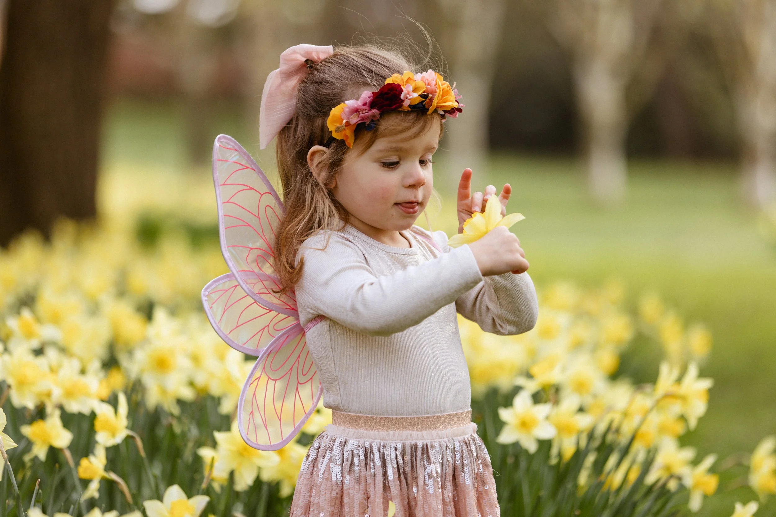 A young girl dressed as a fairy or butterfly, with pink fairy wings, a floral headband, pink skirt with sequins, and a beige top, standing outdoors among yellow daffodils.