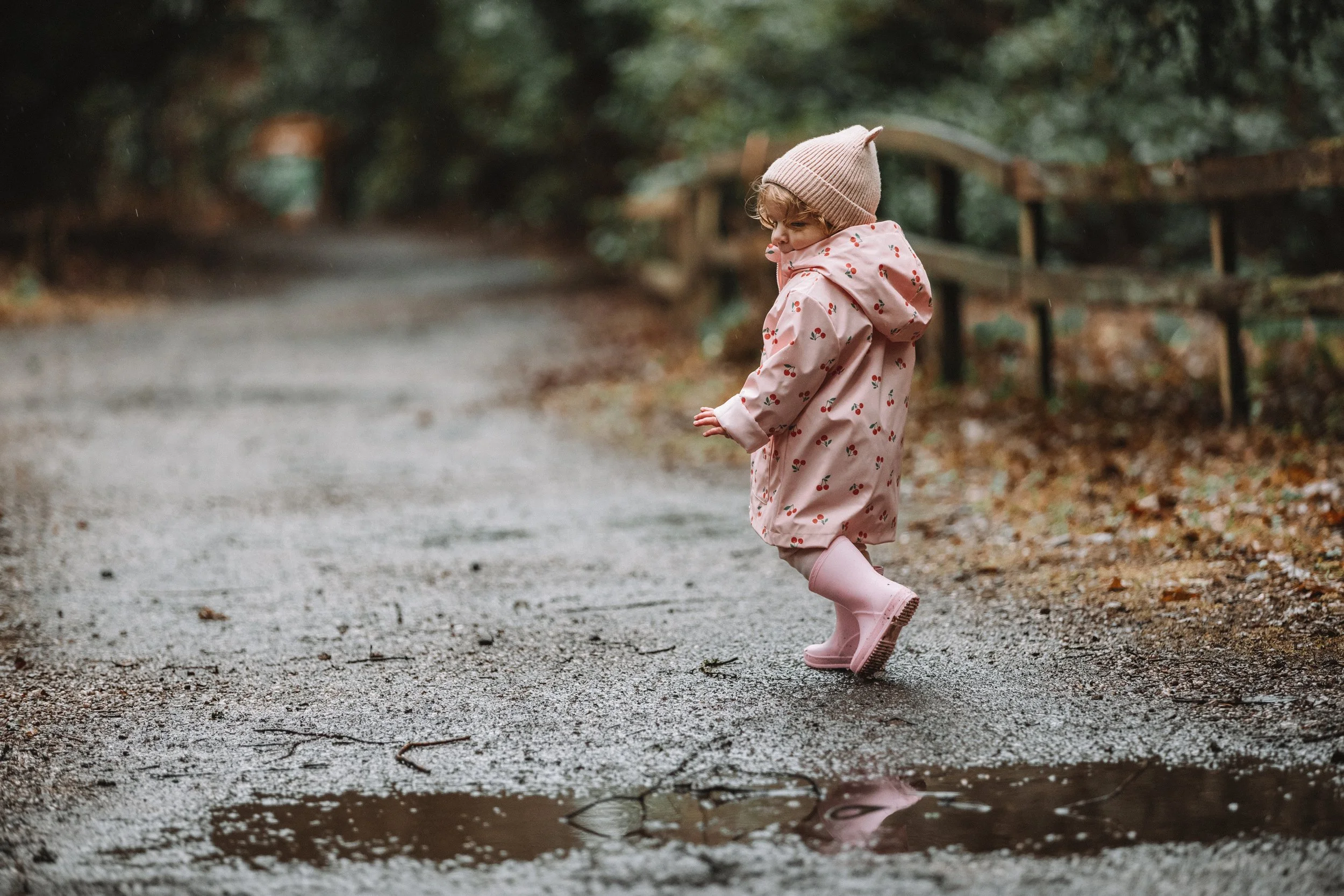 A young girl in pink rain boots and a pink raincoat with a cherry pattern, wearing a beige knit hat, is kicking water in a puddle on a muddy forest path.
