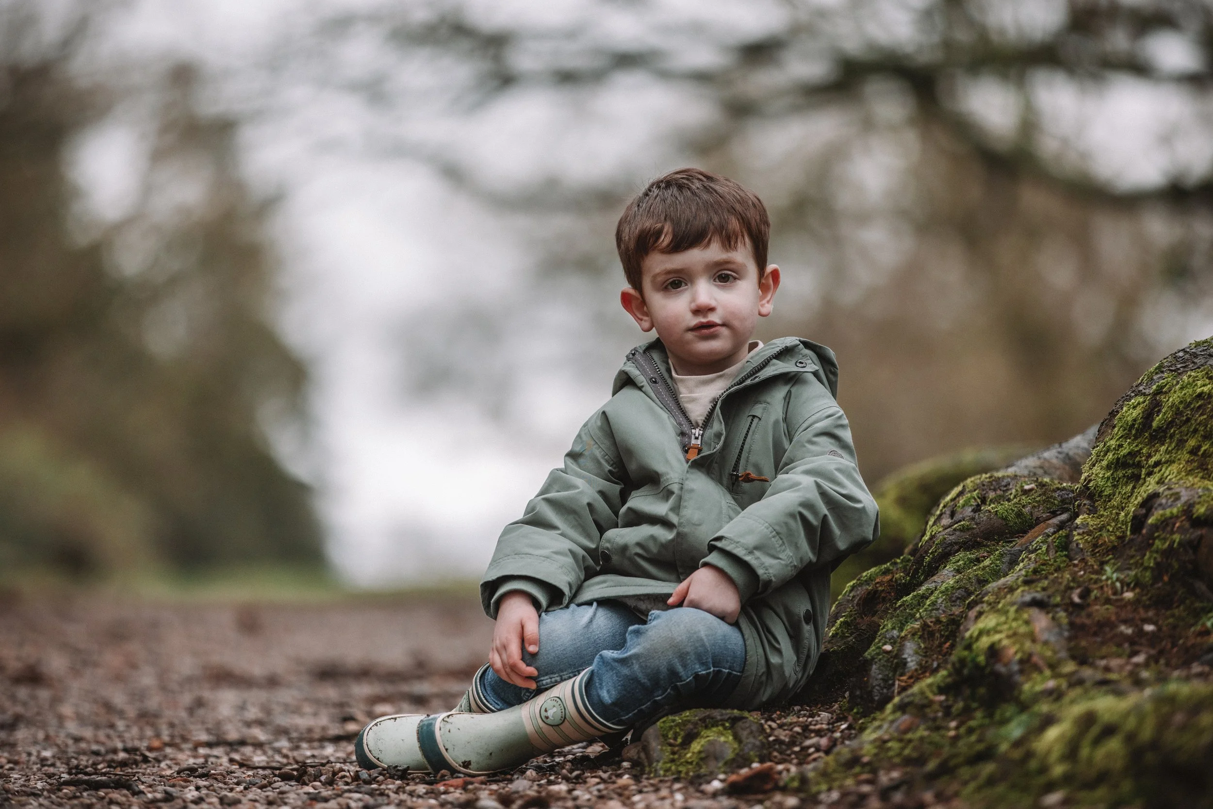 A young boy with brown hair, wearing a green jacket, jeans, and rain boots, sitting on the ground next to moss-covered rocks in a wooded area during daytime.