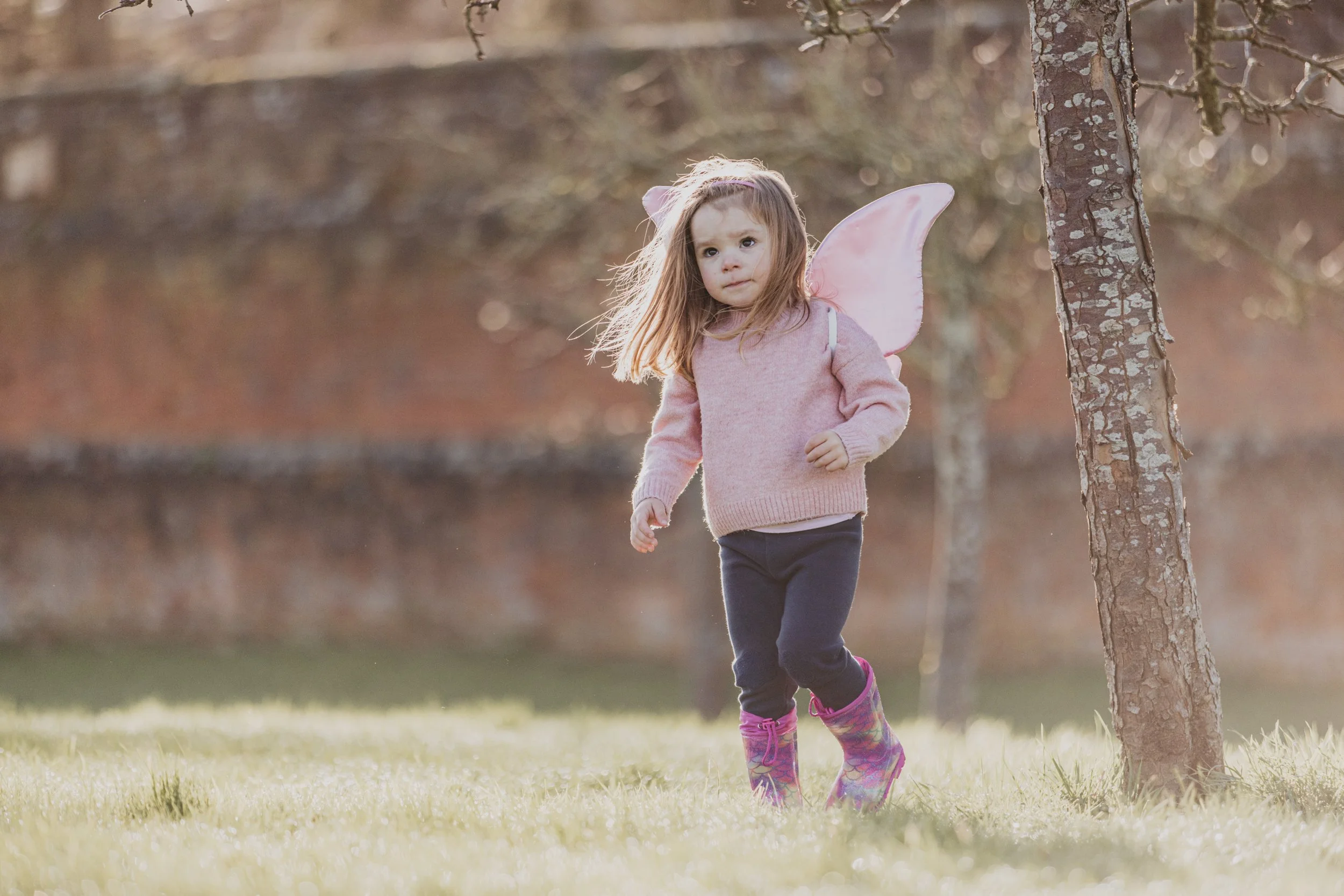 A young girl dressed as a fairy with pink wings, wearing a pink sweater, dark pants, colorful rain boots, walking outdoors beside a tree during daytime.
