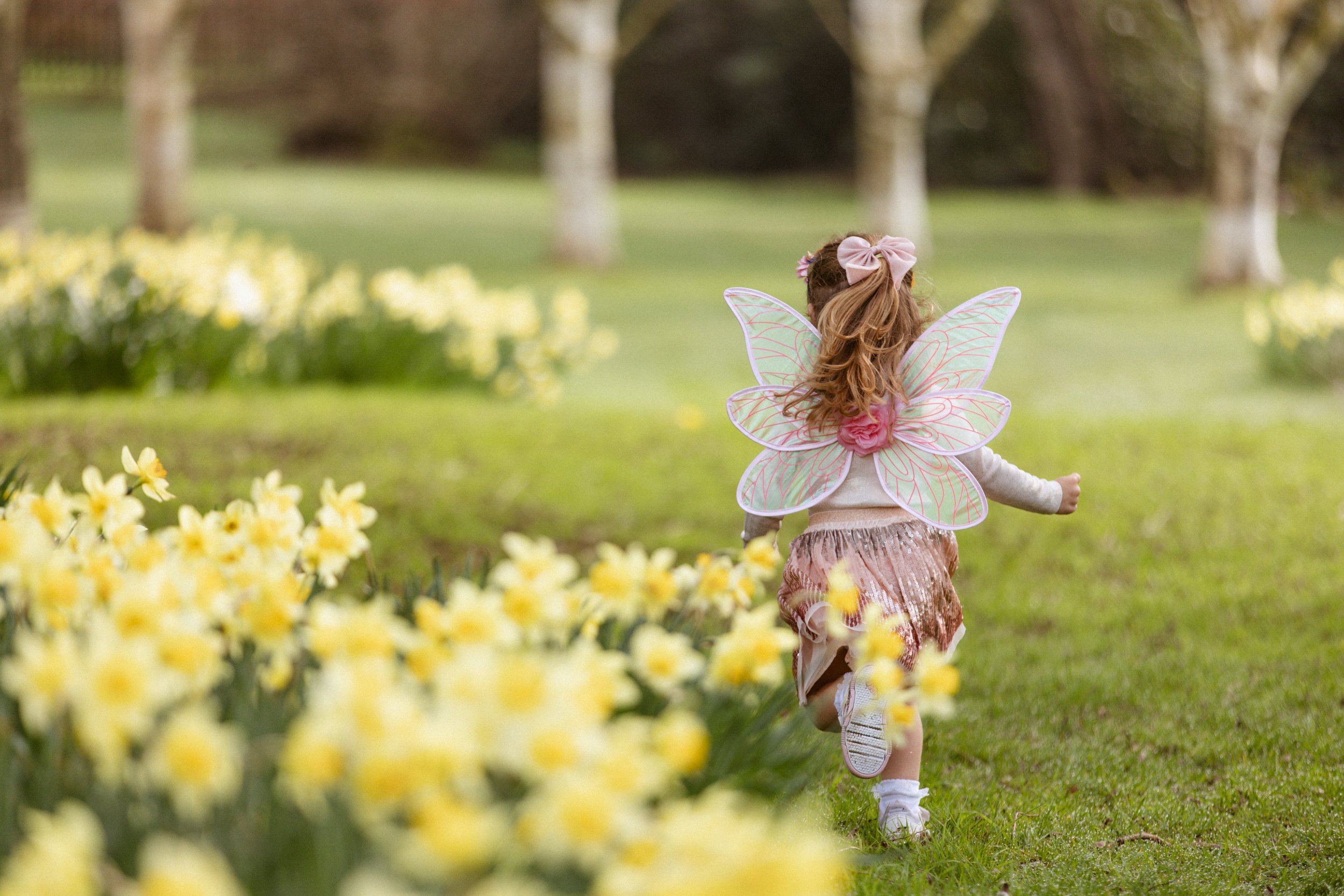 A young girl dressed as a fairy with pink butterfly wings, a pink bow in her hair, and a pink skirt, running through a garden with yellow flowers and trees in the background.
