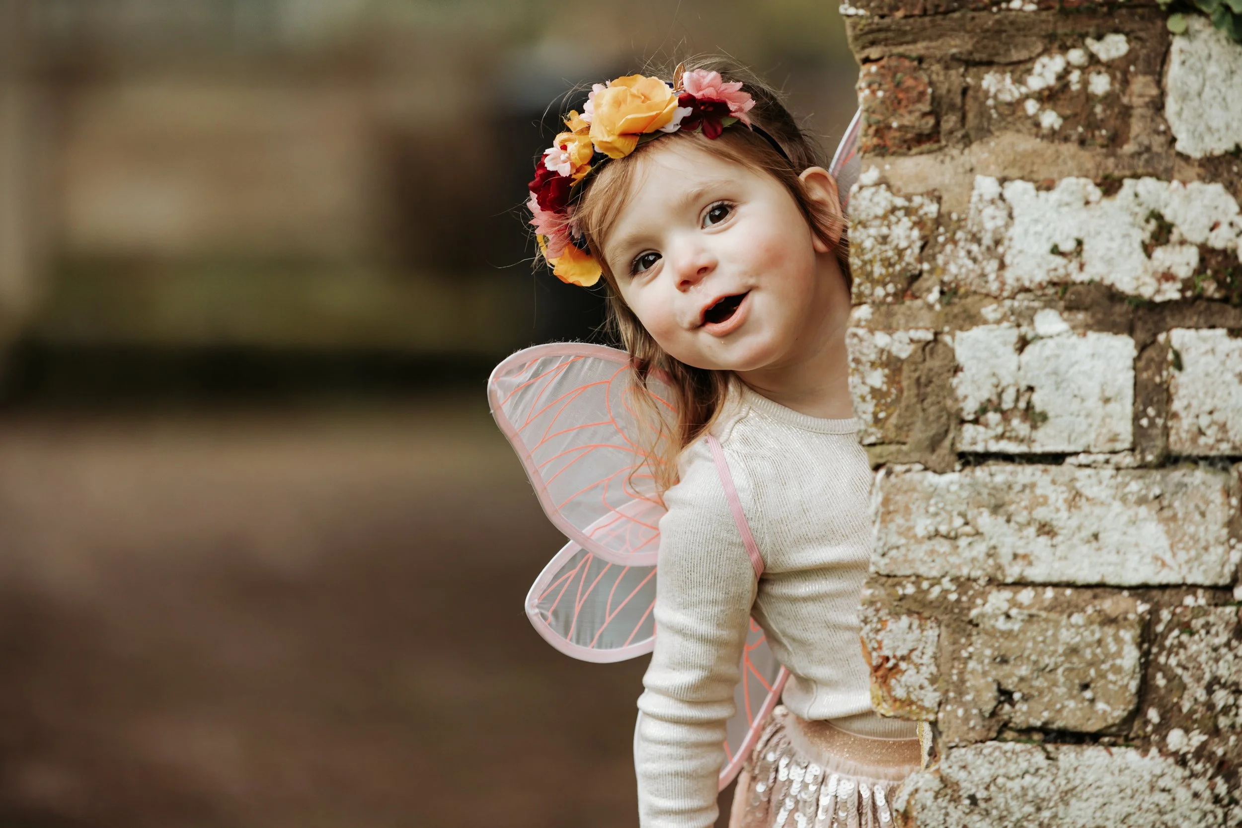 A young girl dressed as a fairy, with pink fairy wings and a colorful flower crown, peeking out from behind a brick wall and smiling.