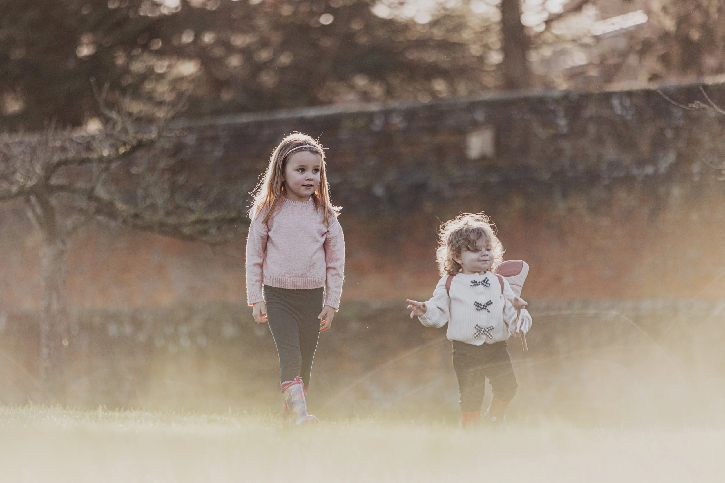 Two young girls walking on a grassy field in autumn, with trees in the background and sunlight filtering through.