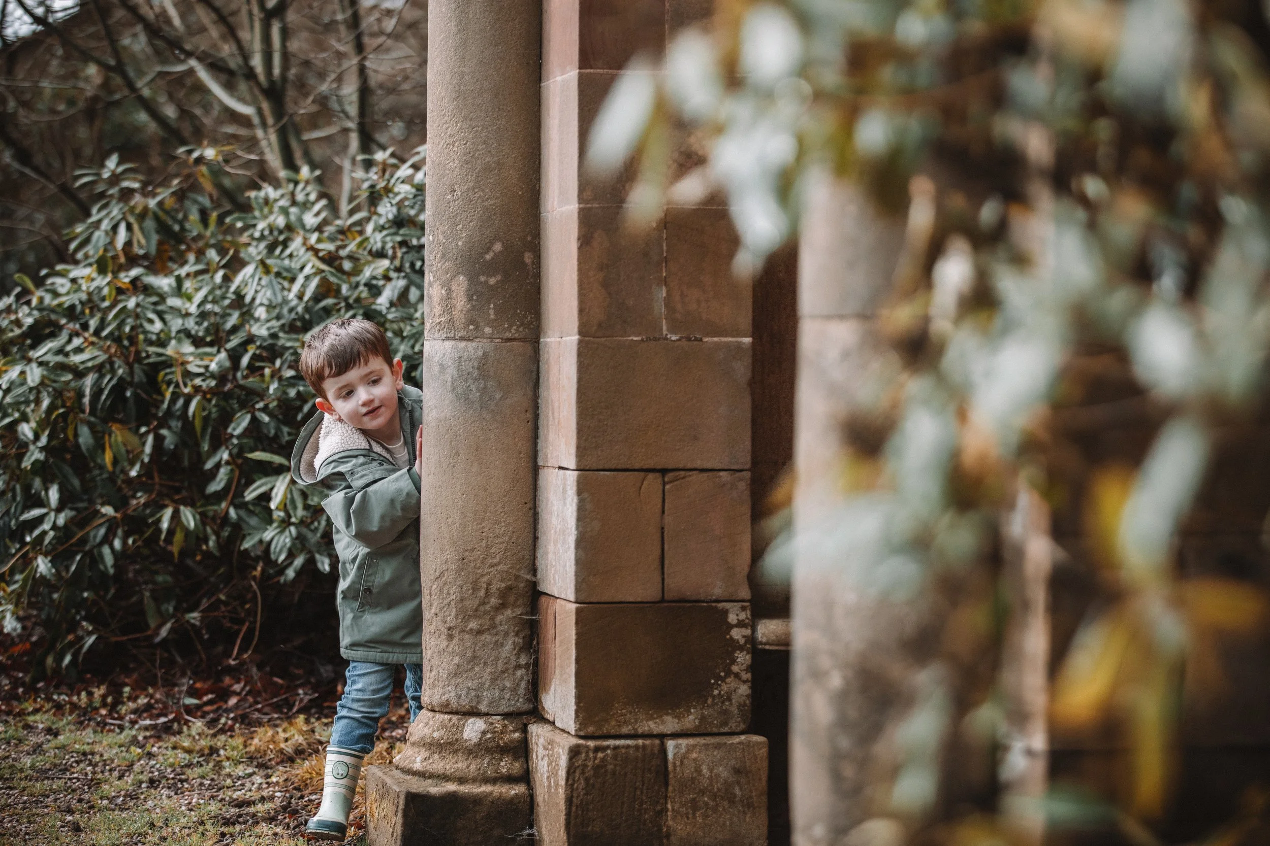 Young boy wearing a green jacket and rain boots peeking from behind a stone pillar in a garden.