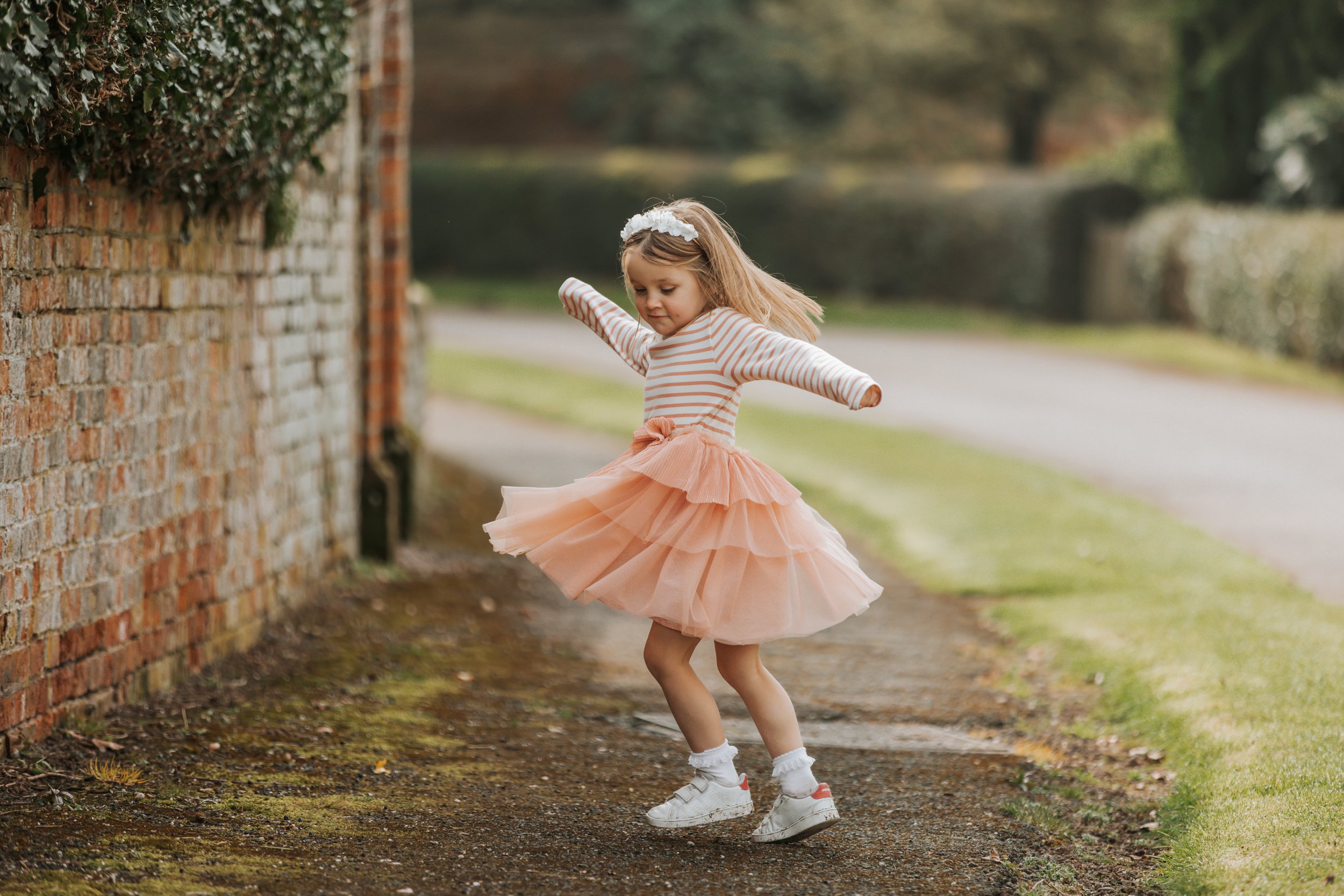 A young girl in a peach tutu skirt, striped long sleeve shirt, white sneakers, and a flower headband spinning on a dirt path next to a brick wall outdoors.