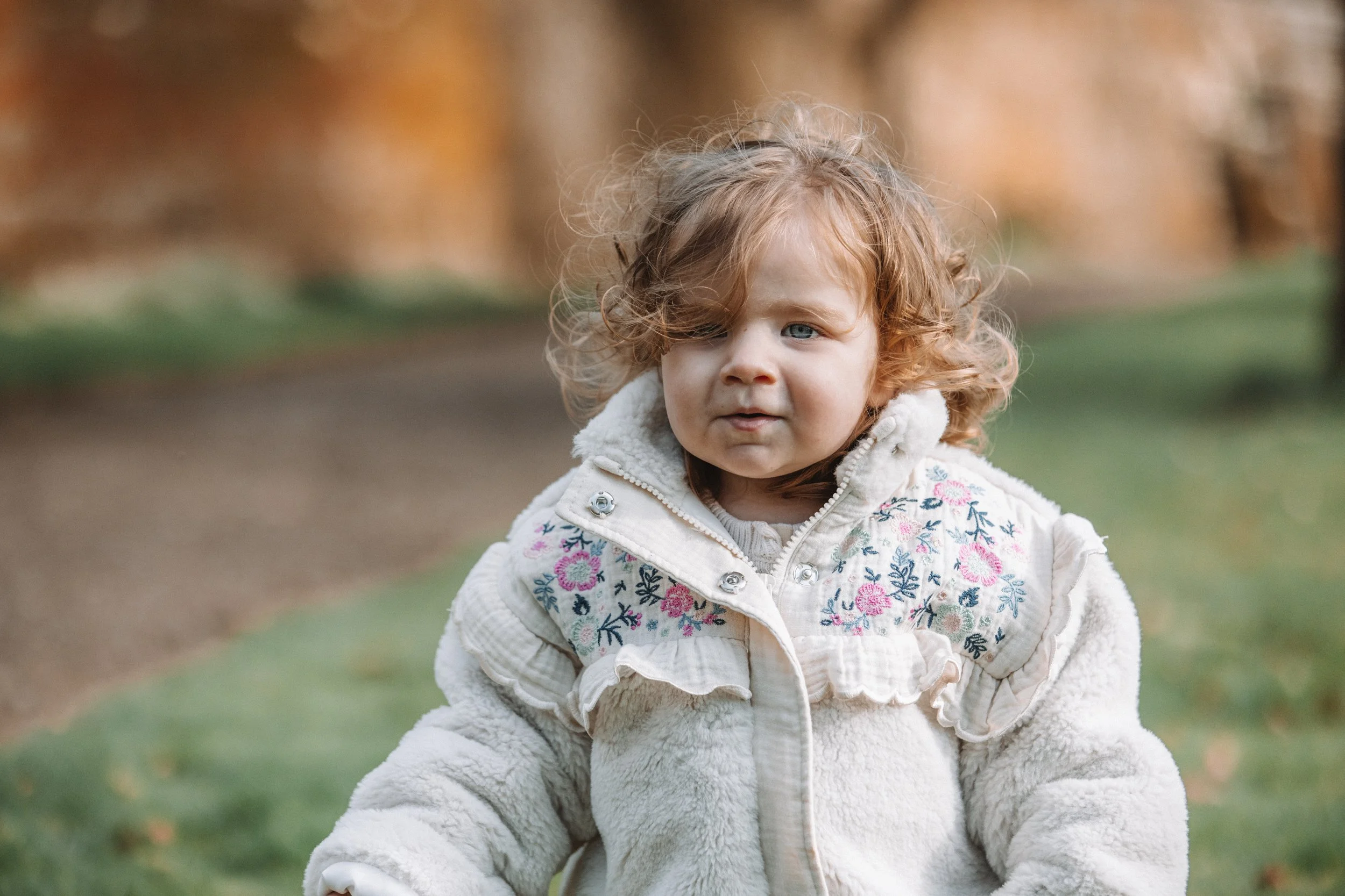 A young girl with curly red hair, wearing a cream-colored jacket with colorful embroidery, standing outdoors in a park with autumn trees in the background.