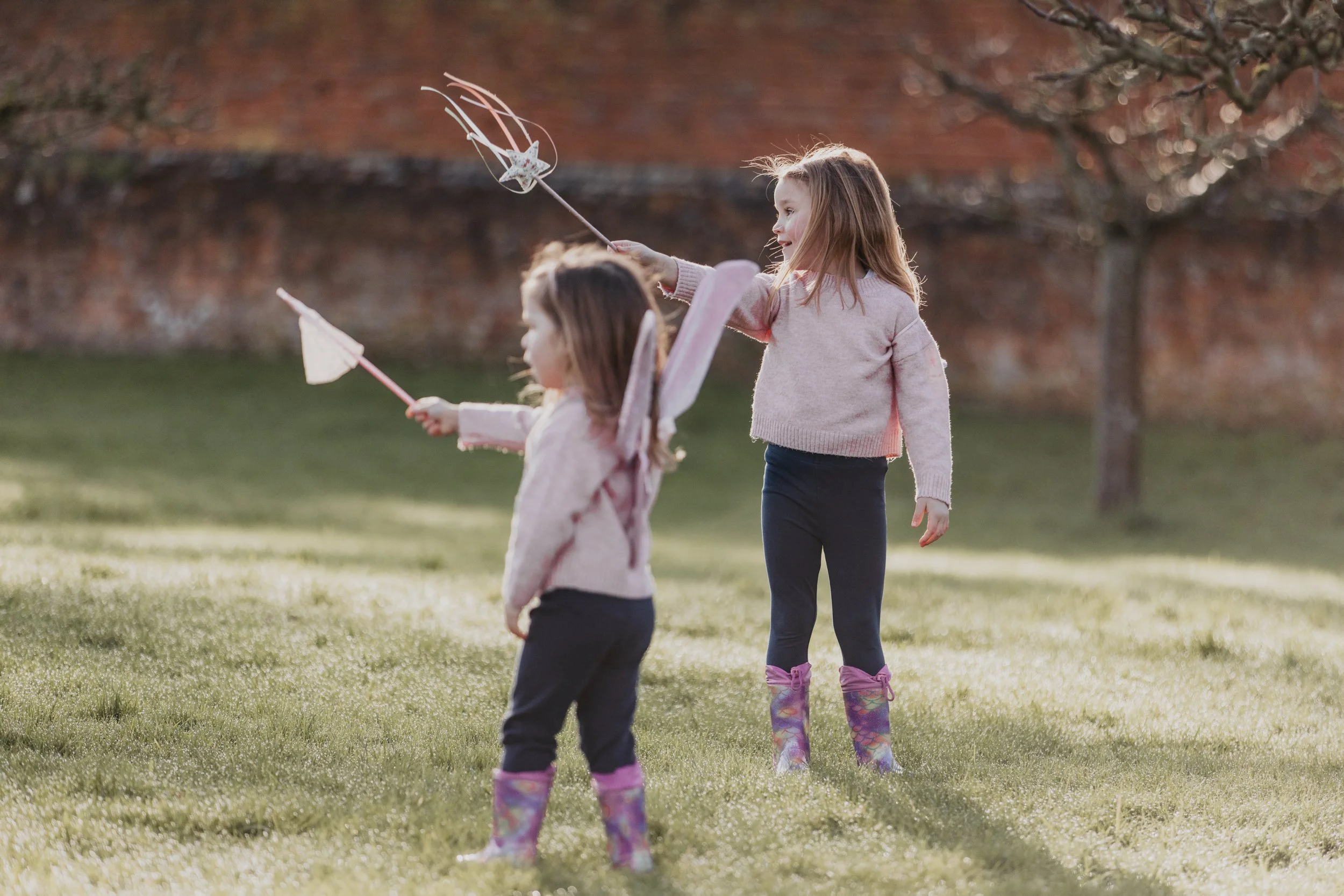 Two young girls in pink sweaters and rain boots playing with fairy wands in a grassy yard during autumn.
