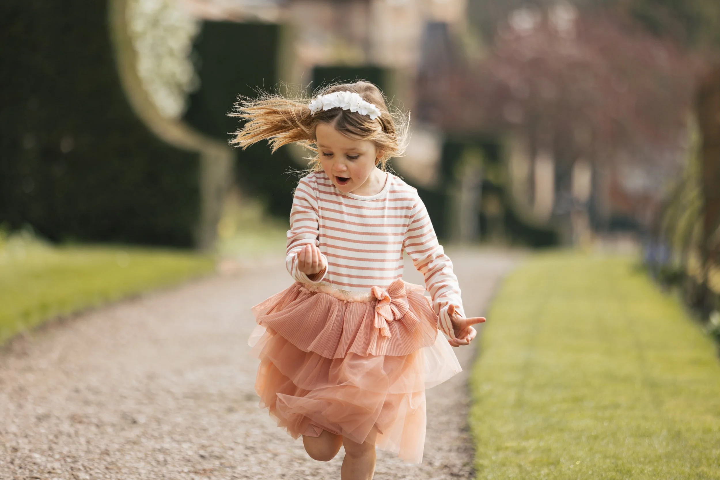 A young girl running on a gravel path in a park, wearing a striped long-sleeve shirt, a peach-colored tutu skirt, and a white floral headband, with her hair flowing and a surprised expression on her face.