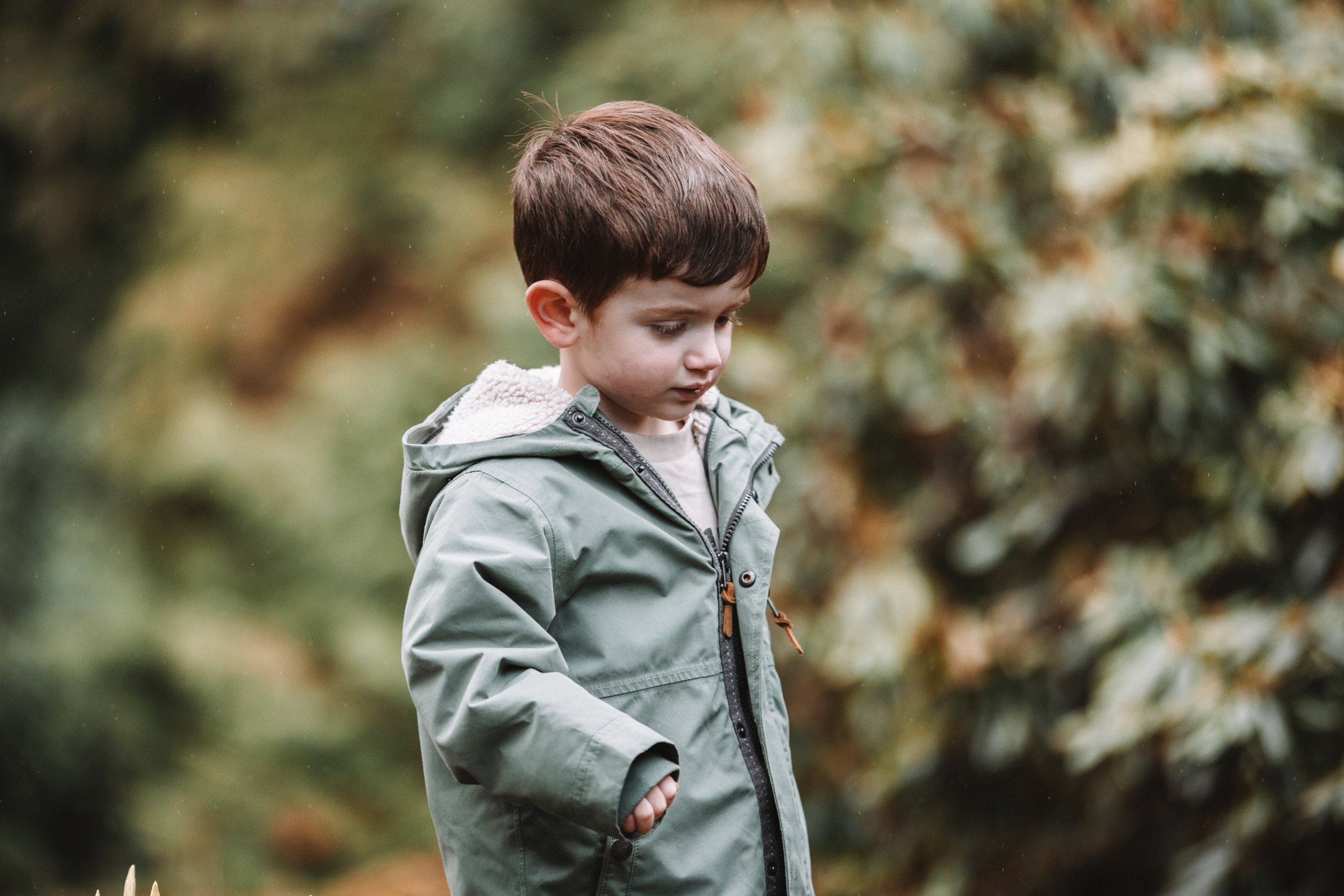 A young boy wearing a gray jacket looking down outdoors with a blurred green and brown background.