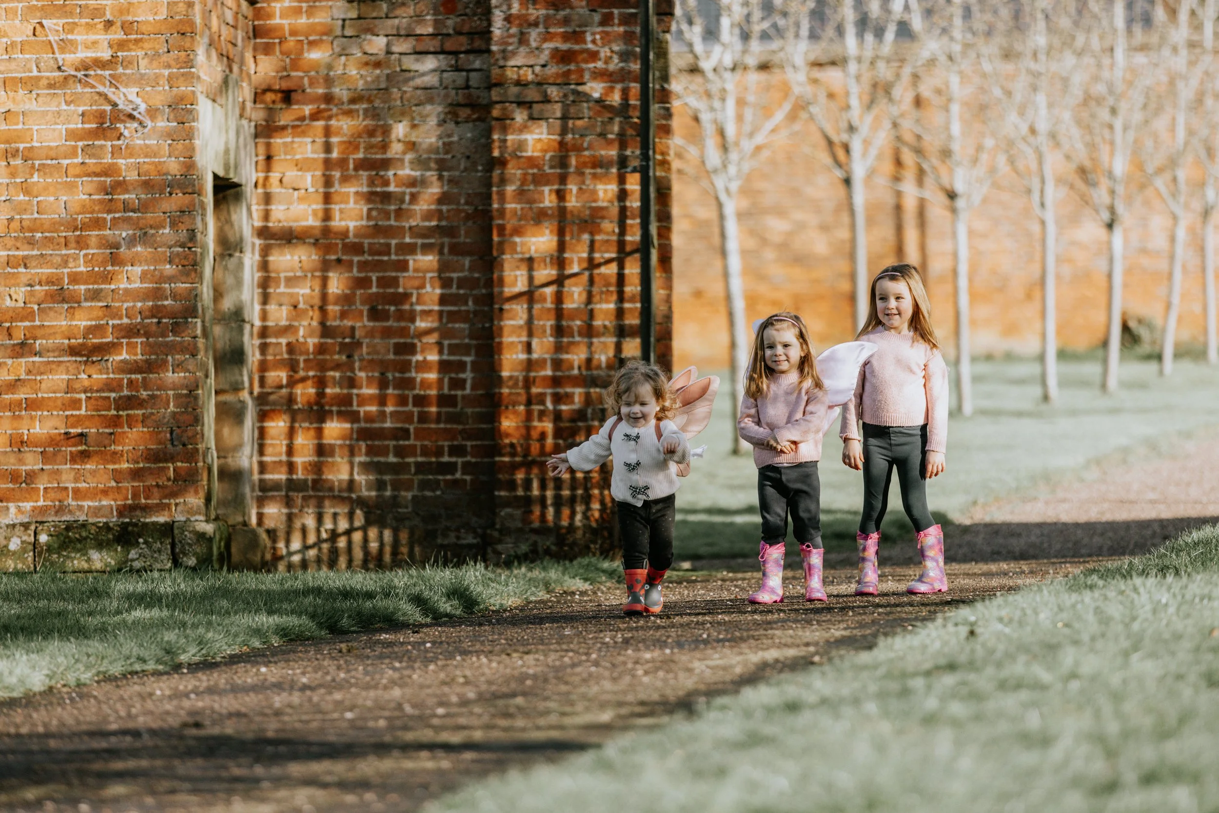 Three young girls walking outdoors near a brick wall and trees in the background, wearing colorful boots and fairy wings.
